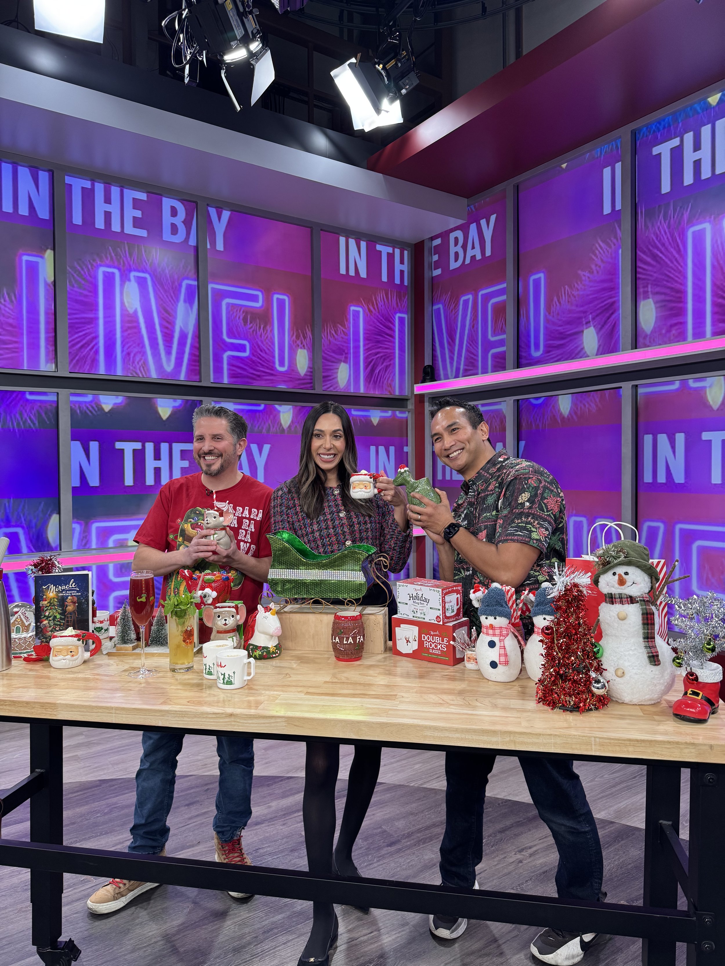 Three people standing behind a table filled with holiday decorations and gifts, smiling and holding holiday-themed mugs, with a colorful holiday display on a background screen.