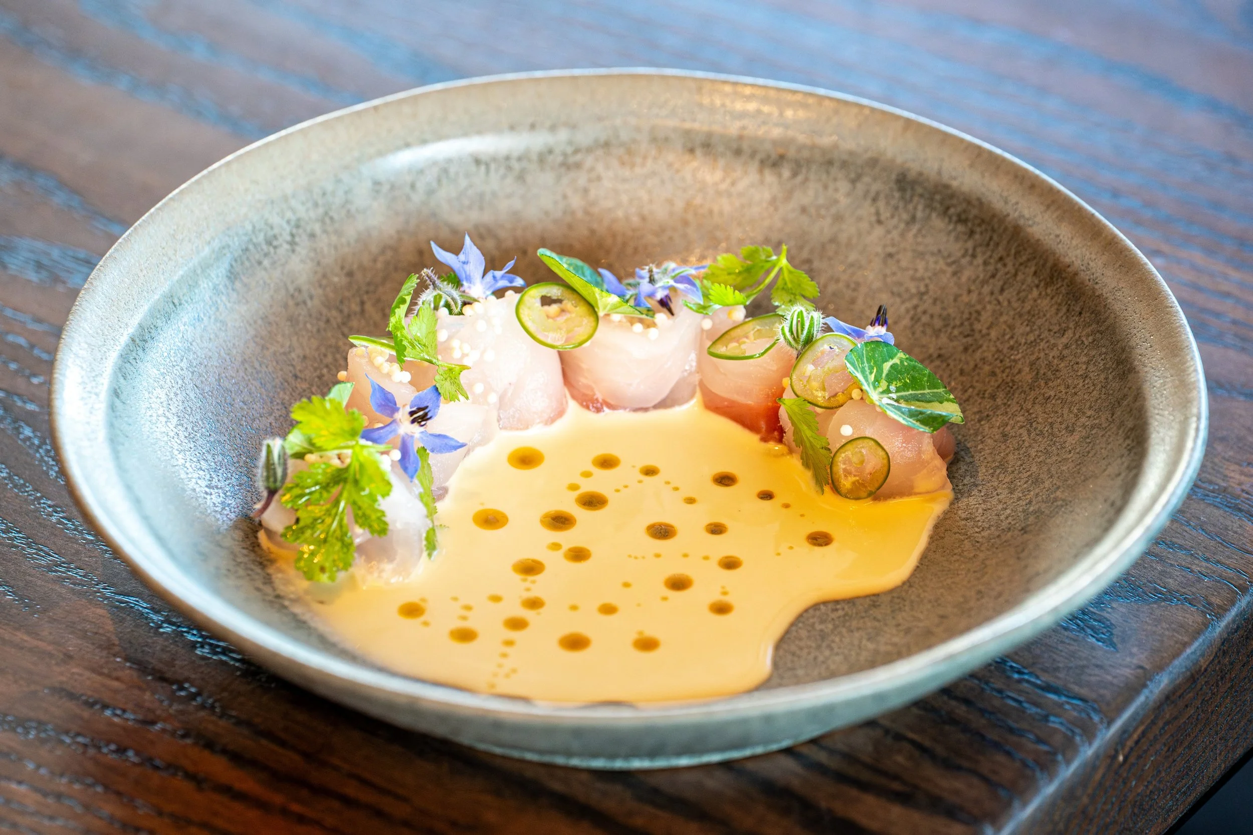 A dish of raw fish, garnished with lime slices, edible flowers, and herbs, served in a yellow sauce with oil droplets, on a dark textured plate.