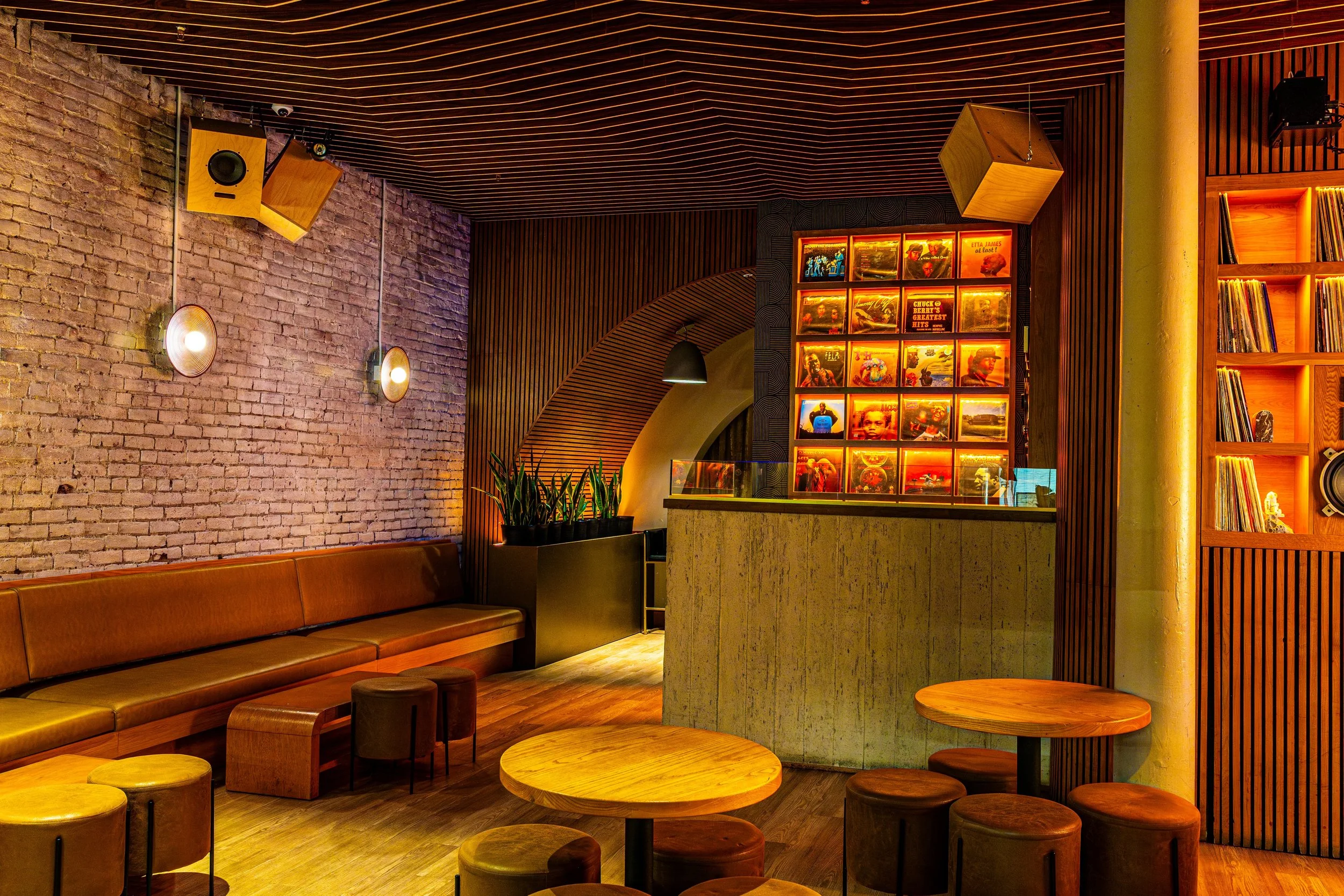 Interior of a lounge or bar with brick walls, wooden ceiling accents, orange lighting, vinyl records on shelves, and wooden tables and stools.