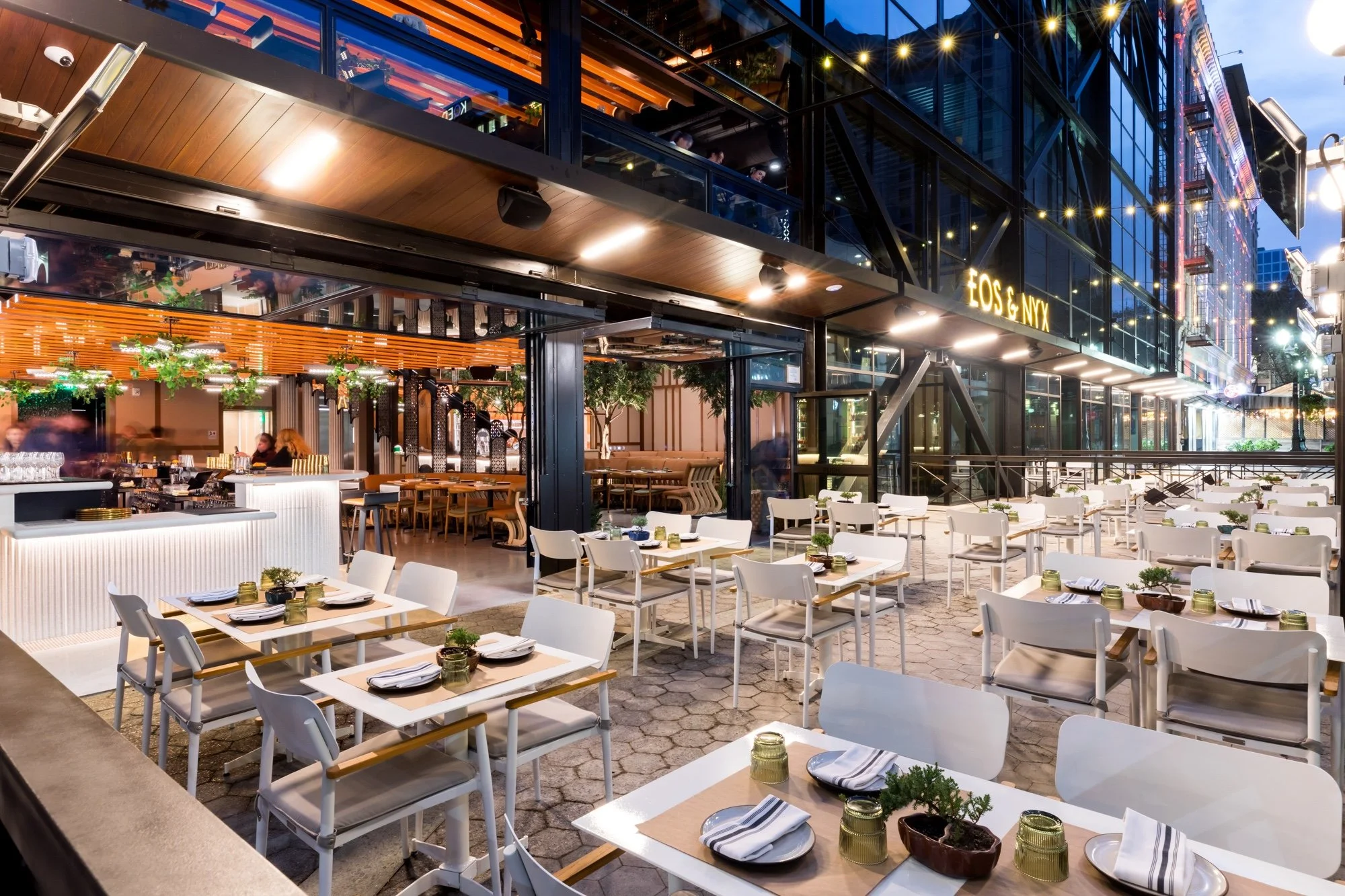 Outdoor dining area with white chairs and tables set with plates and napkins, illuminated by ambient lighting, with a modern building featuring large windows and the sign 'FOOD & NEX' in the background.