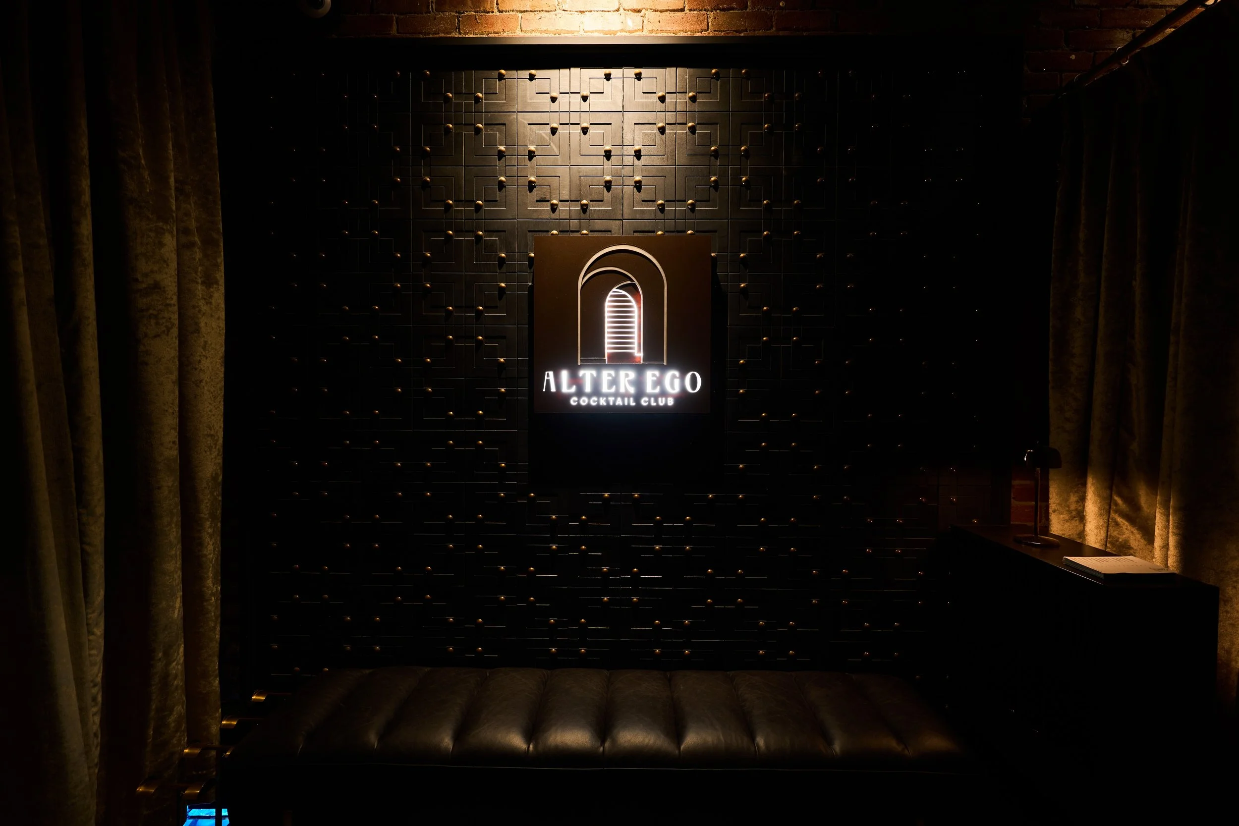 A dark interior of a cocktail club with a black wall featuring a geometric pattern with small round accents. In the center, a lit sign reads 'Alter Ego Cocktail Club' with a logo of an archway. Curtains frame the left and right sides, and there is a 