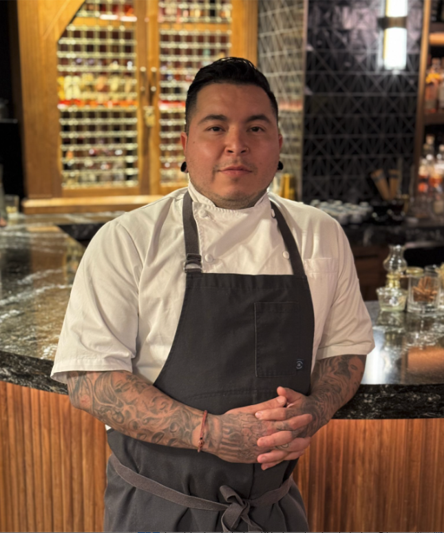 A male chef wearing a white chef's coat and black apron standing in a restaurant or bar with a wooden wine rack and bar counter behind him.