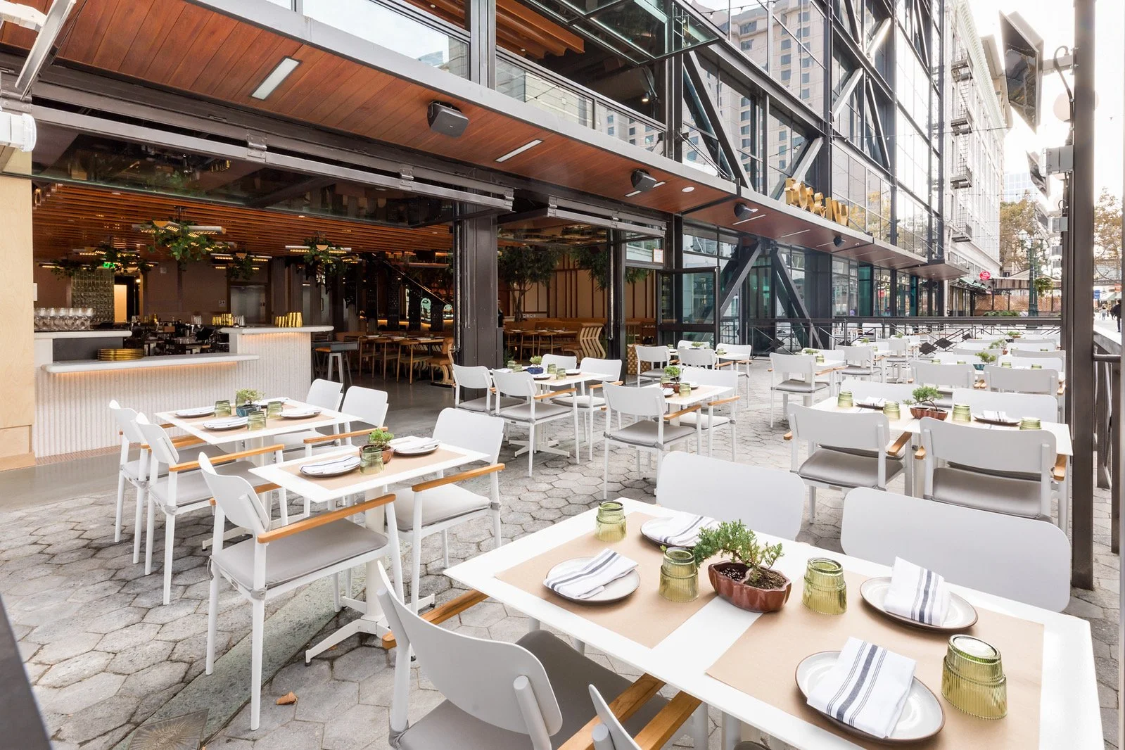 Empty outdoor restaurant patio with white tables and chairs, set for dining with plates, napkins, cups, and small potted plants, adjacent to a modern building with large glass windows.