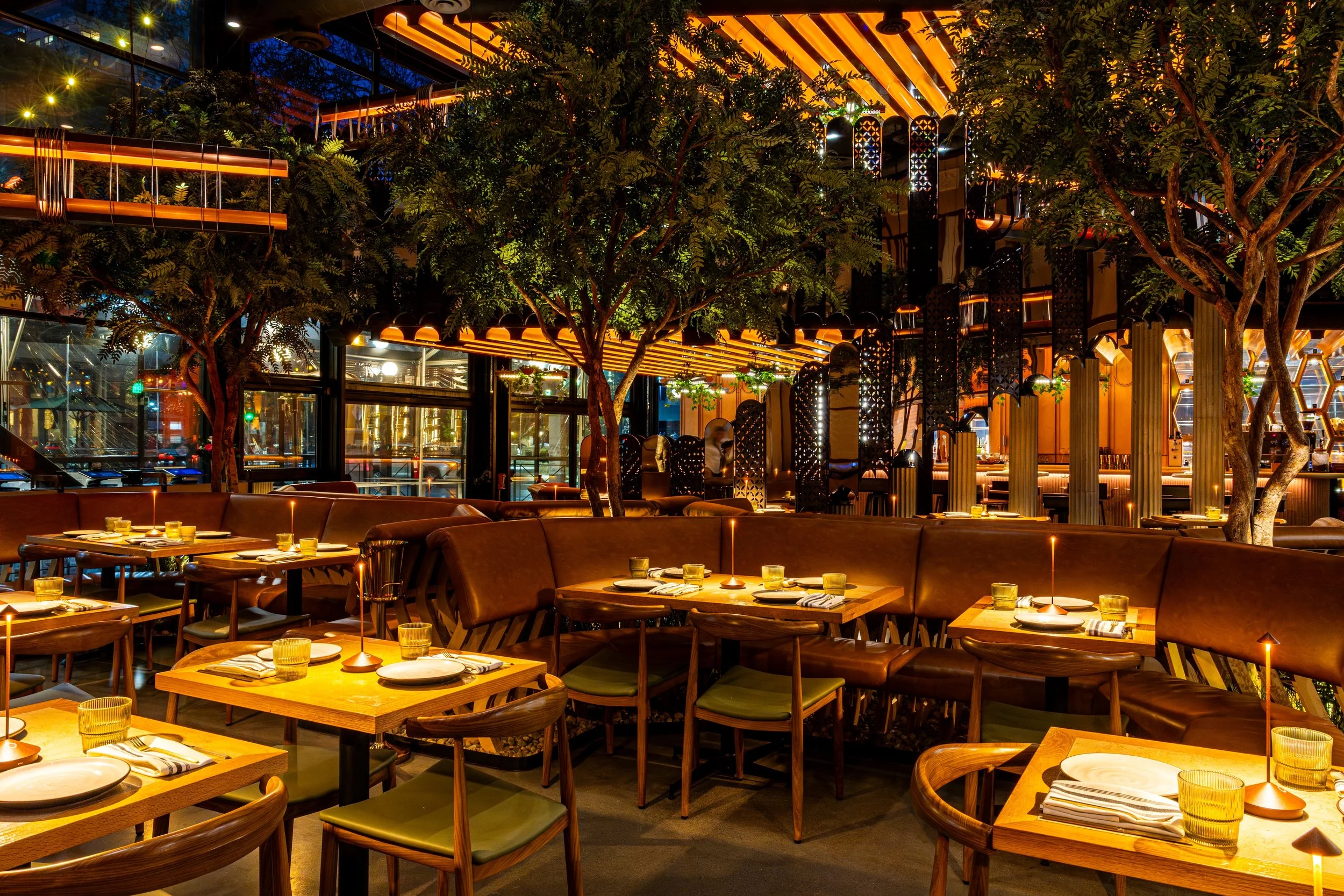 Interior of a modern restaurant with wooden tables set for dining, green upholstered chairs, and decorative lighting. There are lush green trees inside and large glass windows, with a warm orange and black color scheme and ambient lighting.