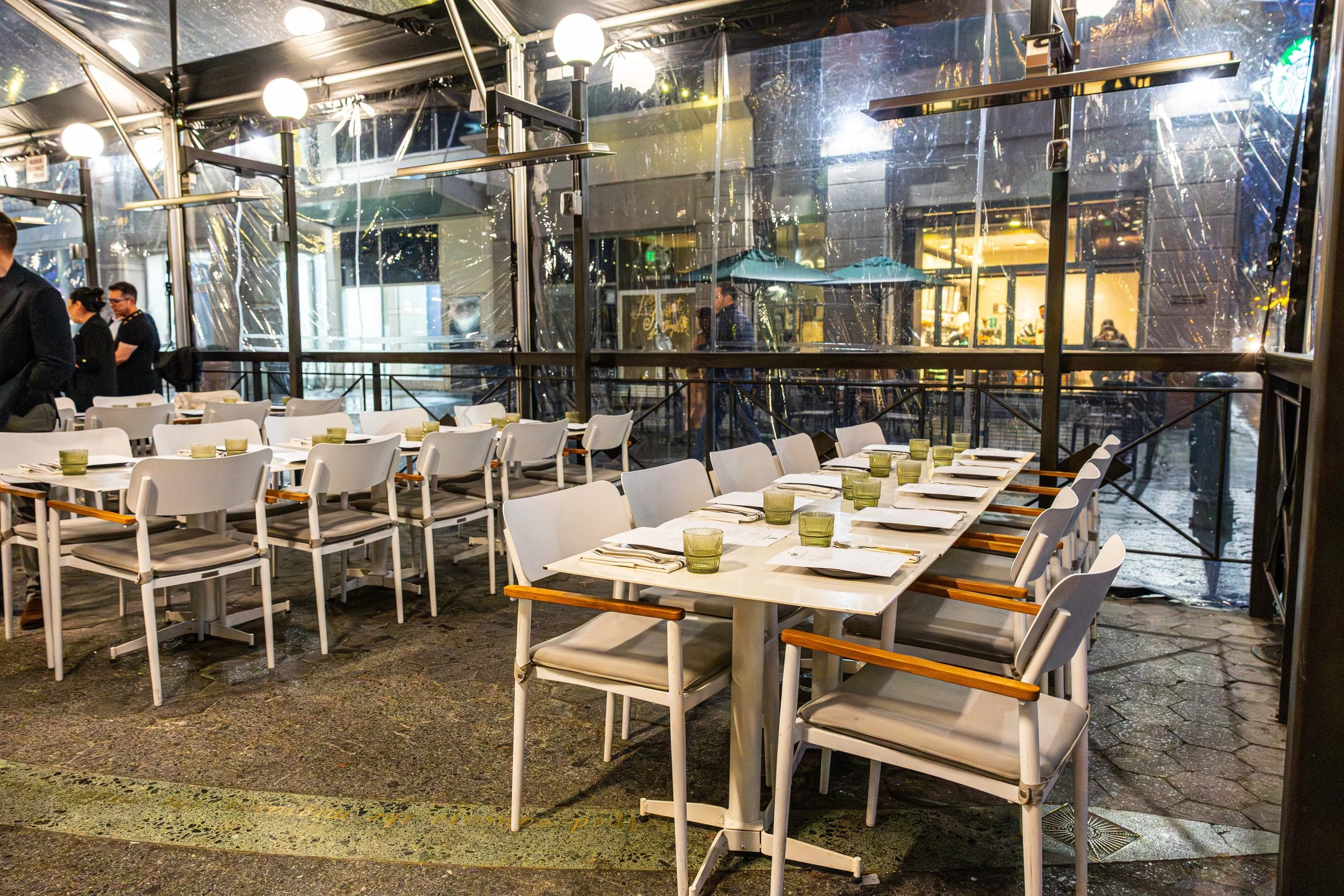 Empty restaurant patio with white chairs around tables set with plates, cutlery, and green glasses, enclosed by transparent plastic panels, with warm indoor lighting and outdoor street scene reflections.