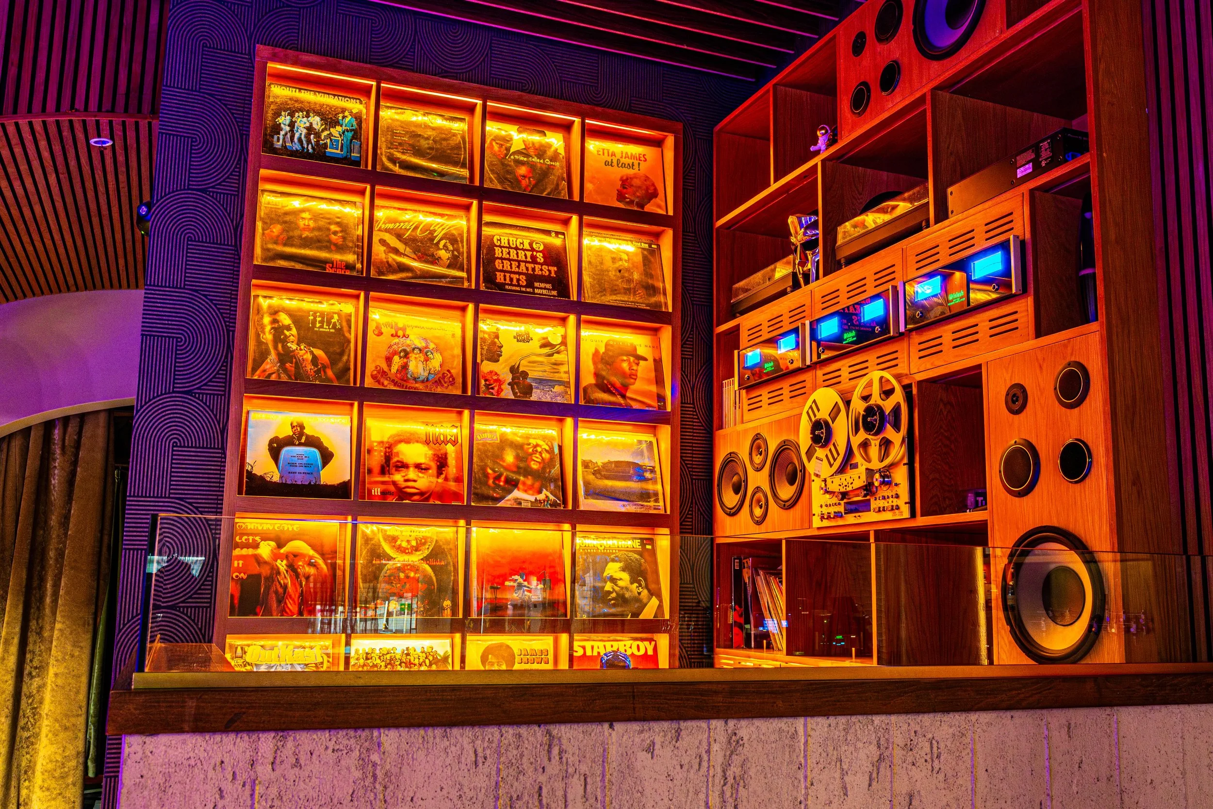 Wall with shelves of vinyl records and audio equipment, illuminated with warm yellow lighting, with a vibrant purple wall and patterned ceiling in the background.