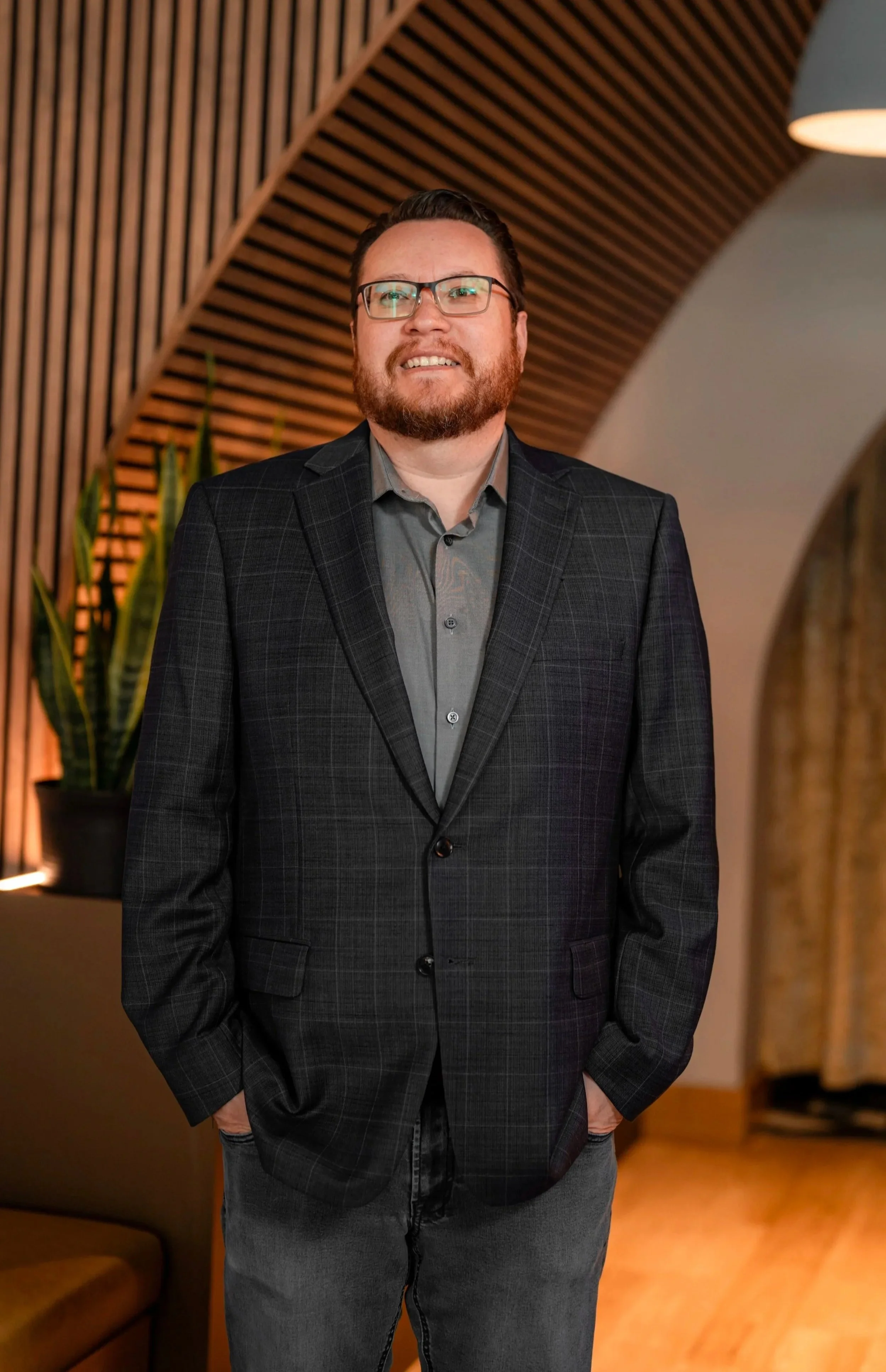 A man with glasses, a beard, and short dark hair wearing a dark checkered blazer over a gray shirt, standing indoors with a wooden, arched ceiling design and plants in the background.