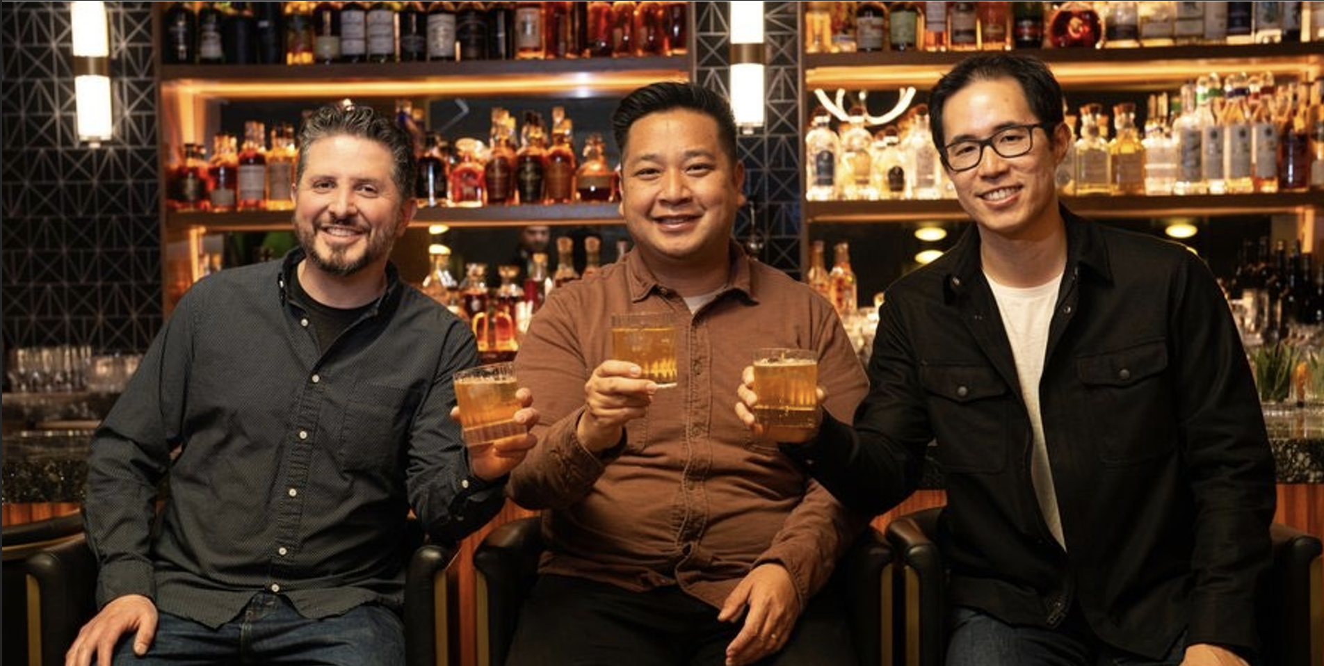 Three men sitting together in a bar, holding drinks, smiling, with shelves of liquor bottles and bar equipment in the background.