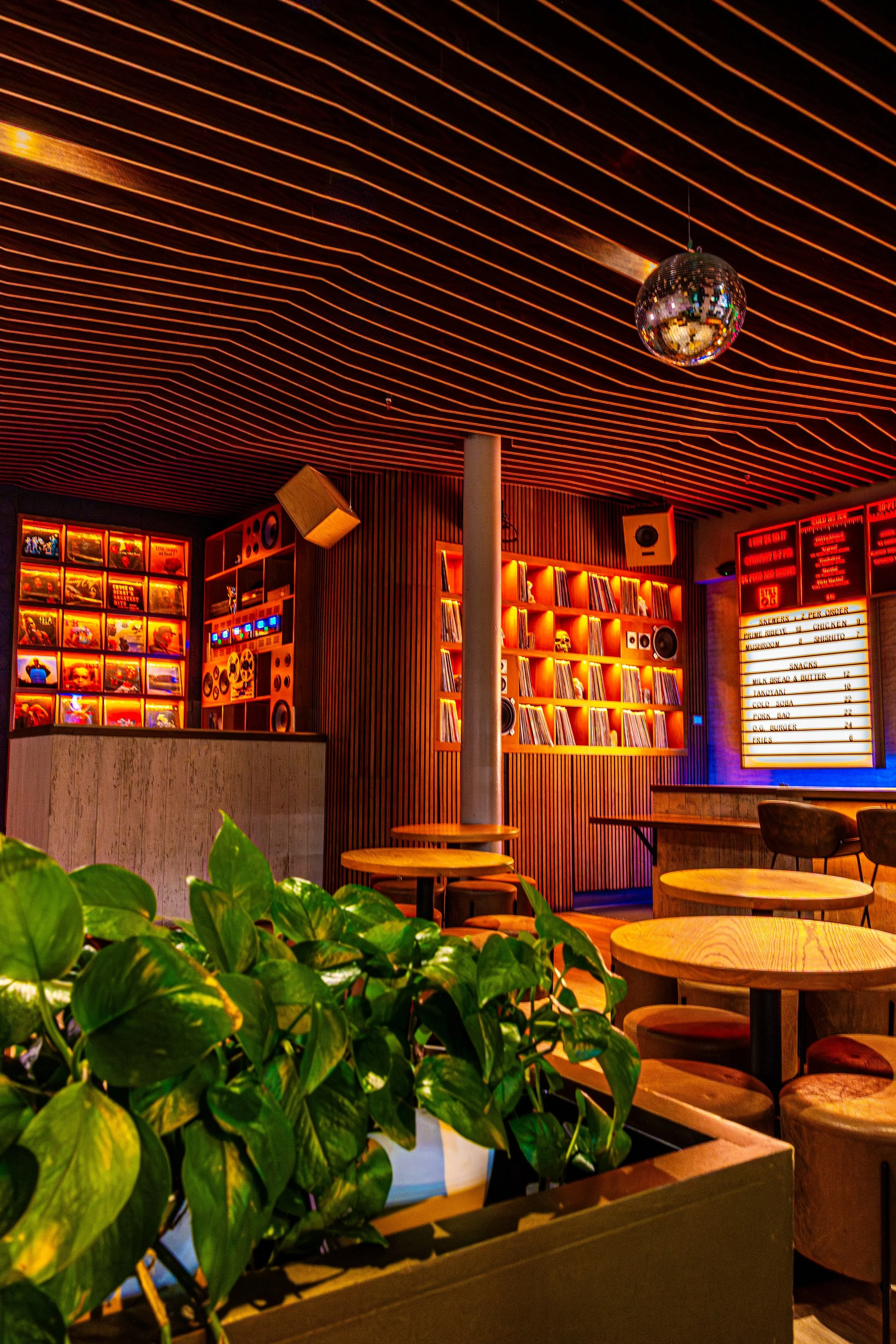 Interior of a cozy bar or cafe with wooden tables and chairs, neon lighting, wall shelves with books, vinyl records, and a large menu board. A disco ball hangs from the ceiling, and there are green plants in the foreground.