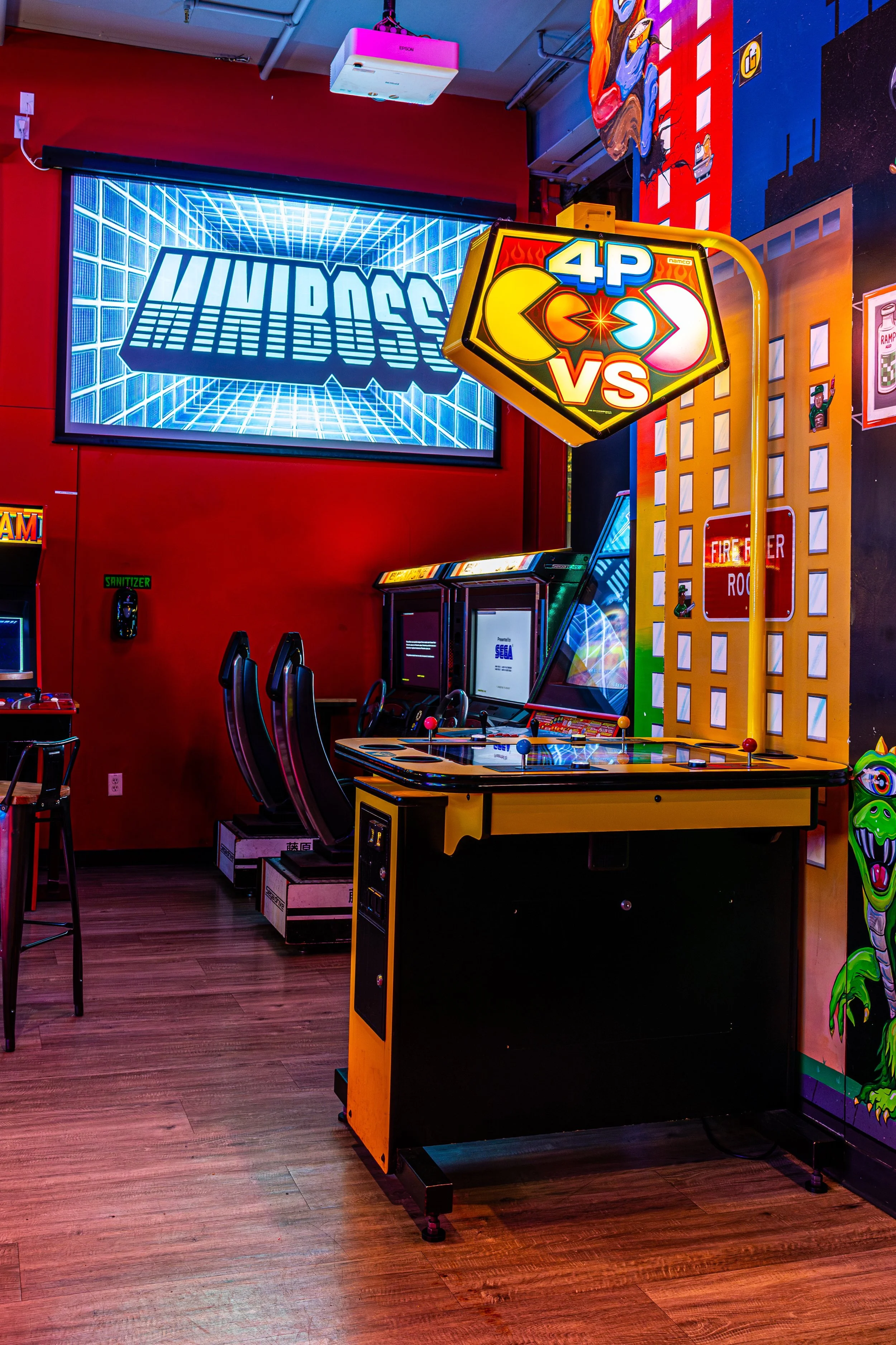 An arcade gaming room with a classic arcade machine and a Pac-Man themed pinball machine, colorful walls, and a large neon 'MiniBoss' sign.