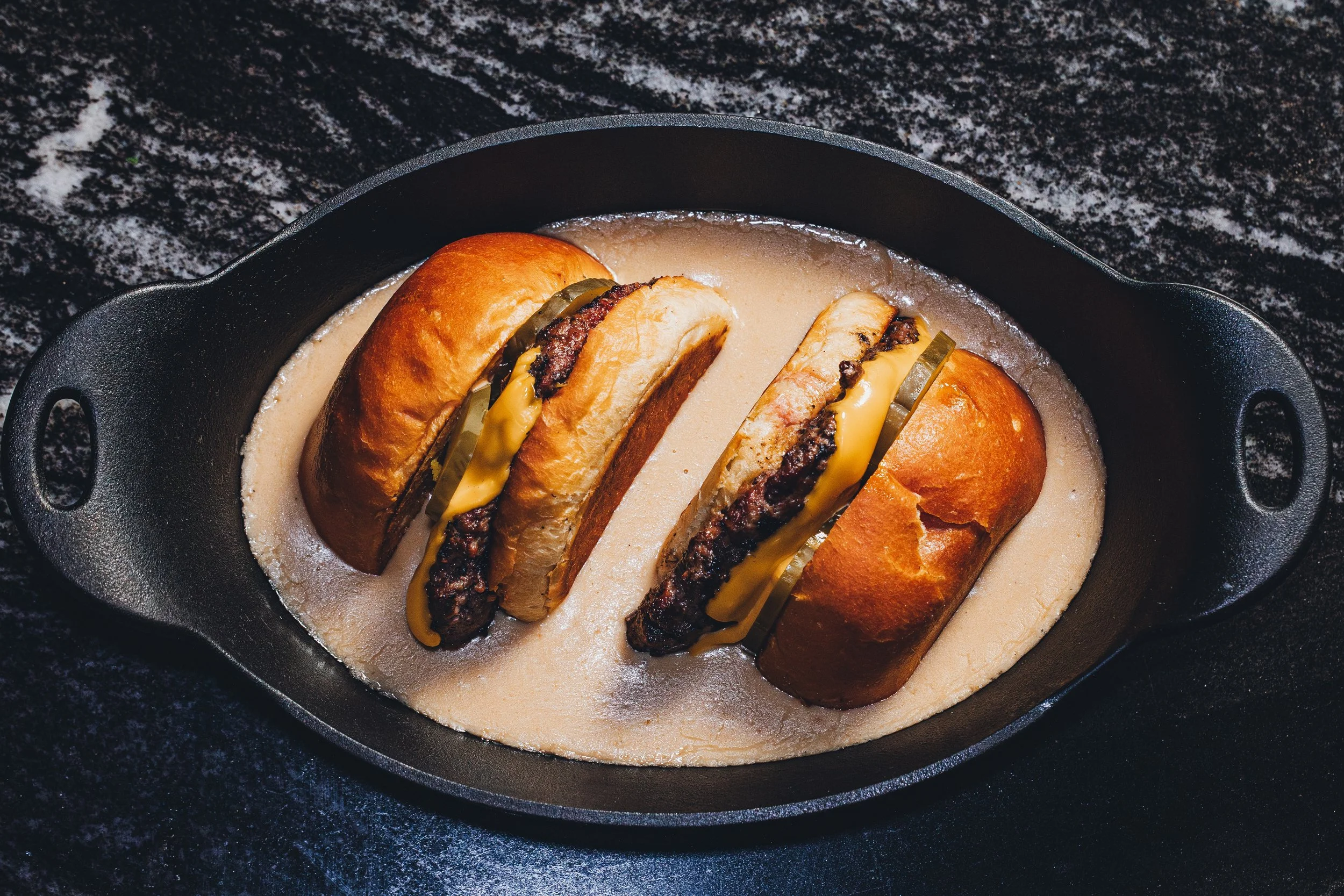 Cast iron skillet containing a hot beef sandwich with melted cheese and pickles on a toasted bun, served with gravy on a dark speckled surface.