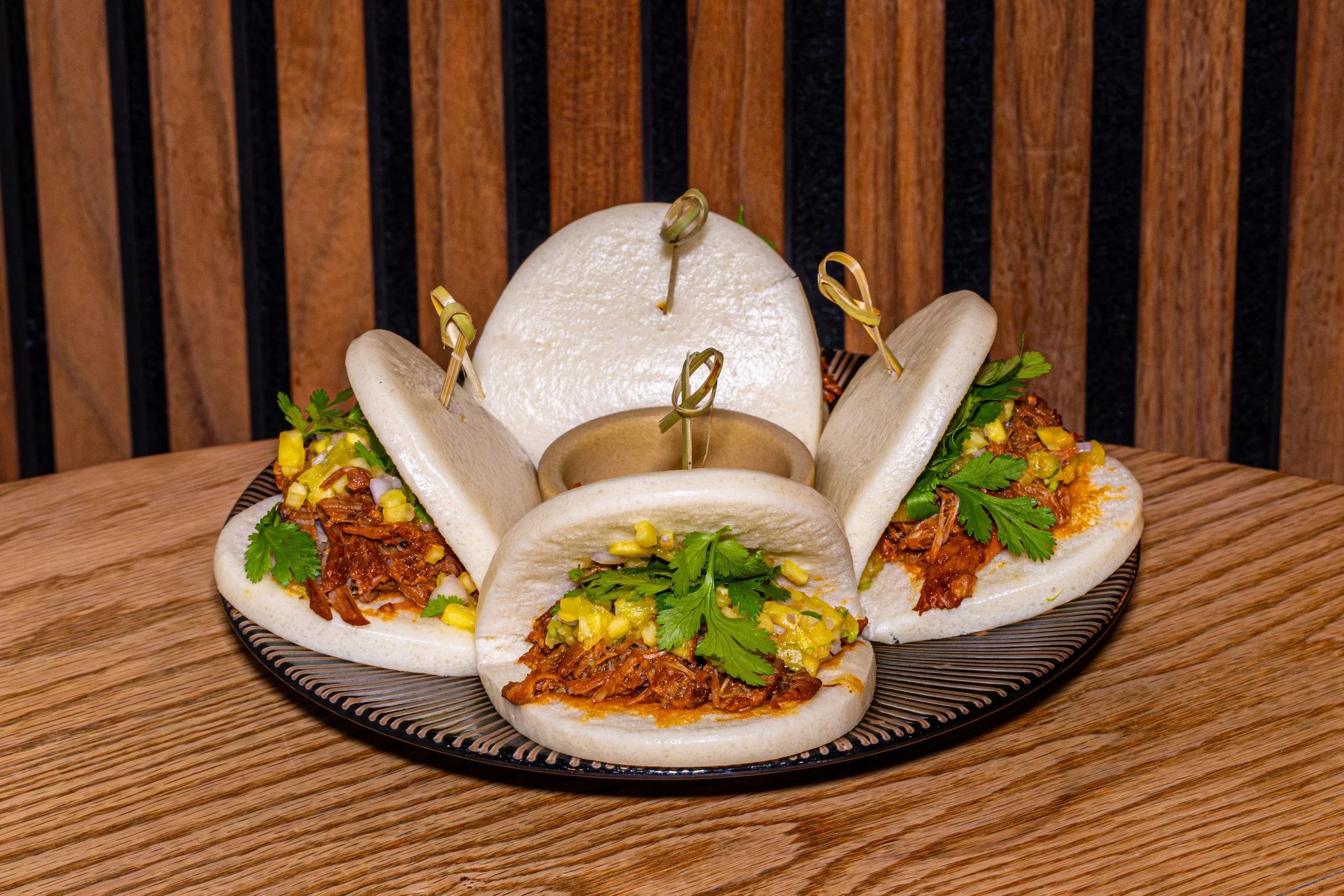 A plate of Mexican tacos filled with shredded meat, garnished with cilantro and chopped onions, with a large white onion in the center and skewers inserted into it, set on a wooden surface with a wooden background.