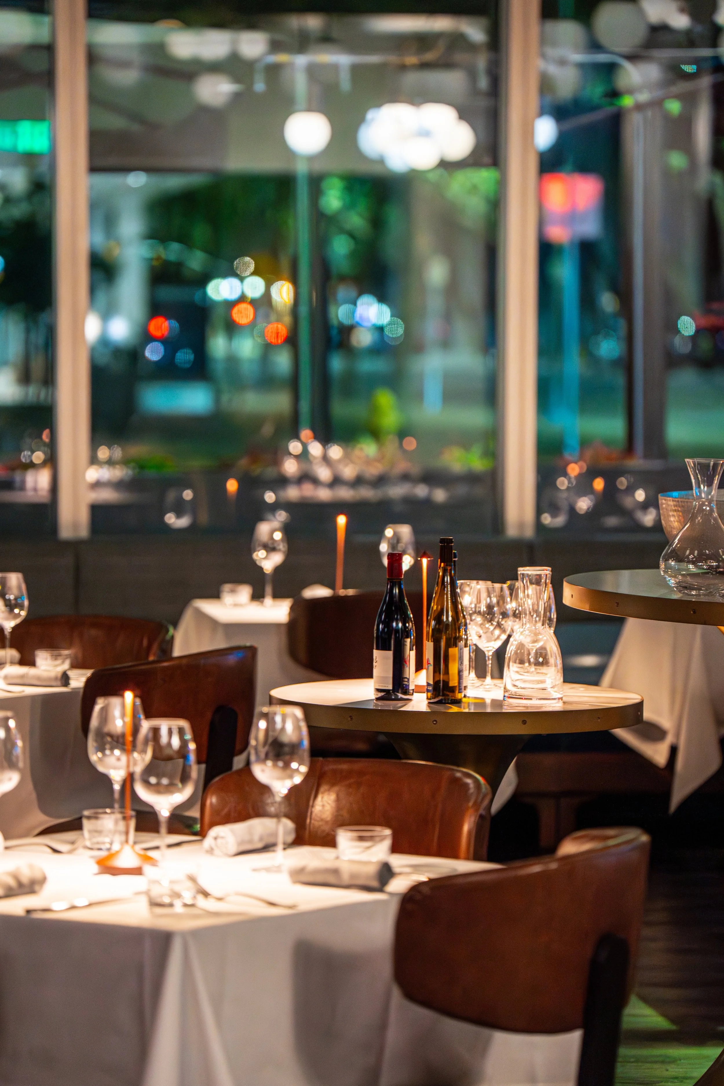 Empty restaurant tables set with white tablecloths, wine glasses, and bottles with a window in the background showing city lights and trees at night.
