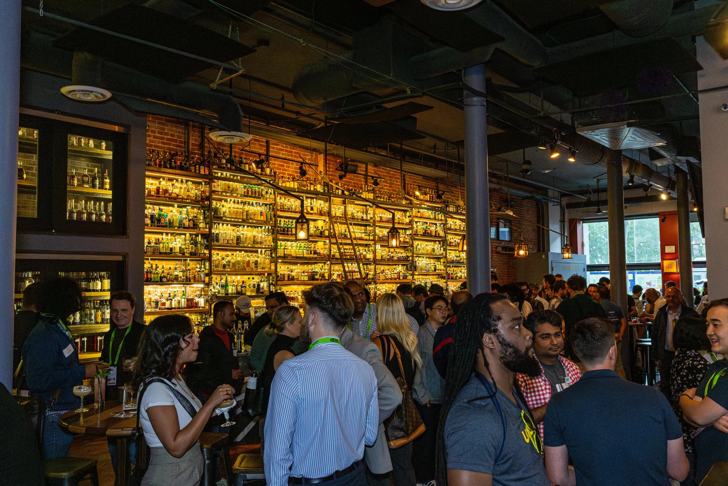 A crowded bar with a large selection of liquor bottles displayed on illuminated shelves against a brick wall. People are gathered, socializing and enjoying drinks inside a modern, industrial-style venue with black ceiling panels and exposed ductwork.
