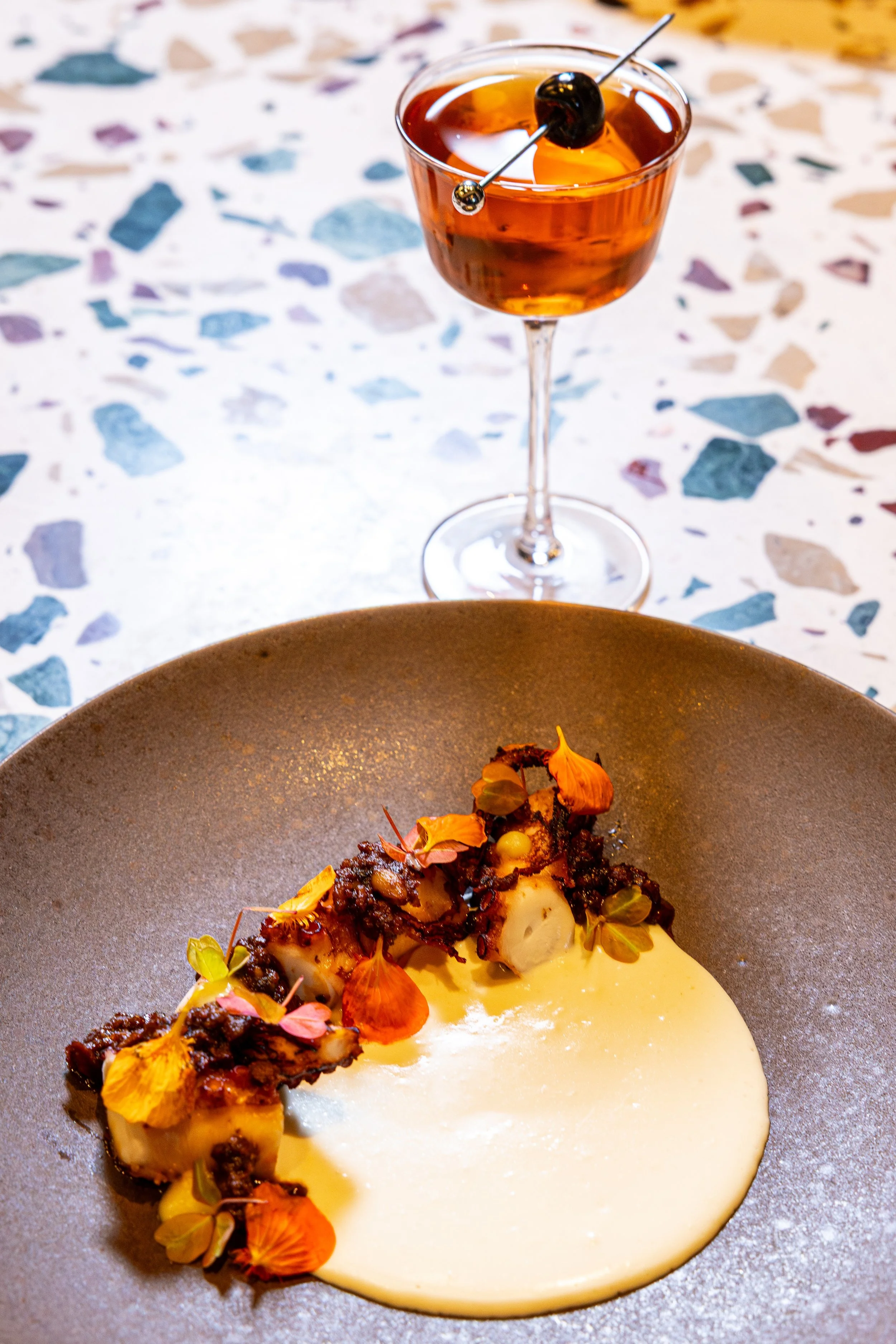 A plated gourmet dish with a roasted vegetable medley topped with edible flowers, served with a light sauce, and a cocktail with a cherry garnish on a terrazzo table.
