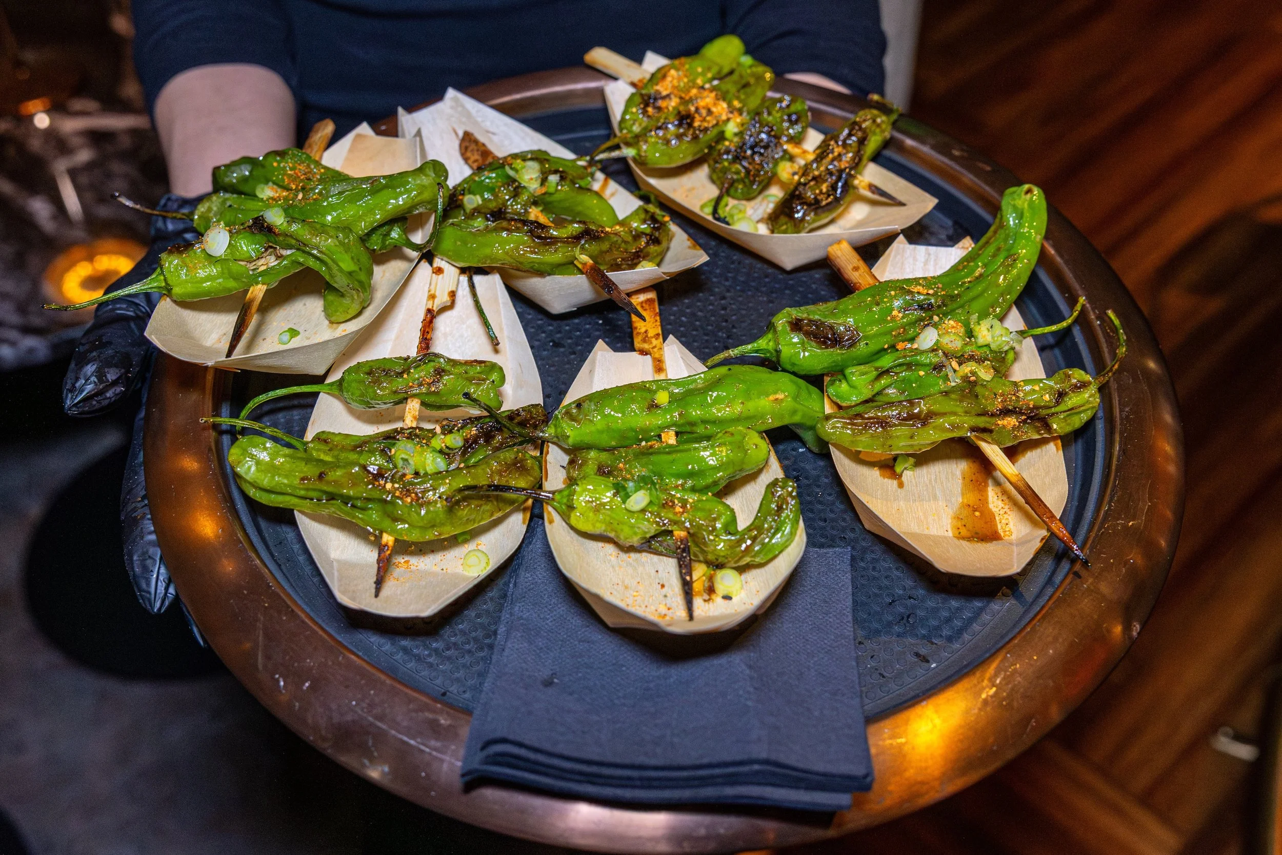Grilled green peppers served on skewers on a tray lined with paper, with some green onions sprinkled on top.