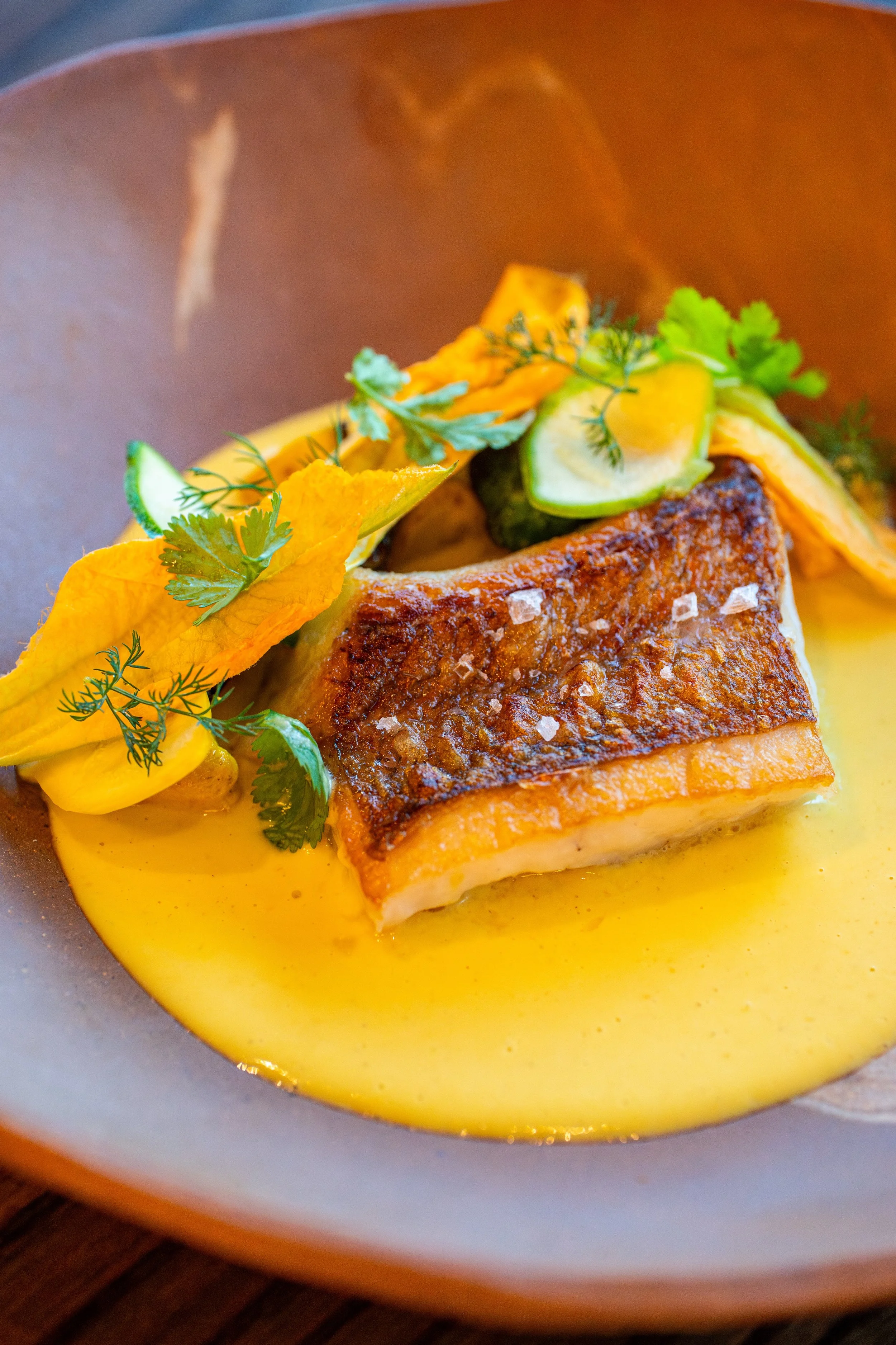 Seared fish fillet served on a bed of creamy yellow sauce with garnishes of fresh herbs and sliced cucumber.
