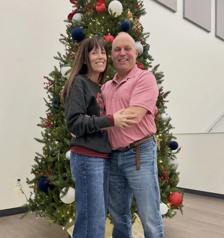 A smiling couple hugging in front of a decorated Christmas tree with red, white, blue, and gold ornaments.