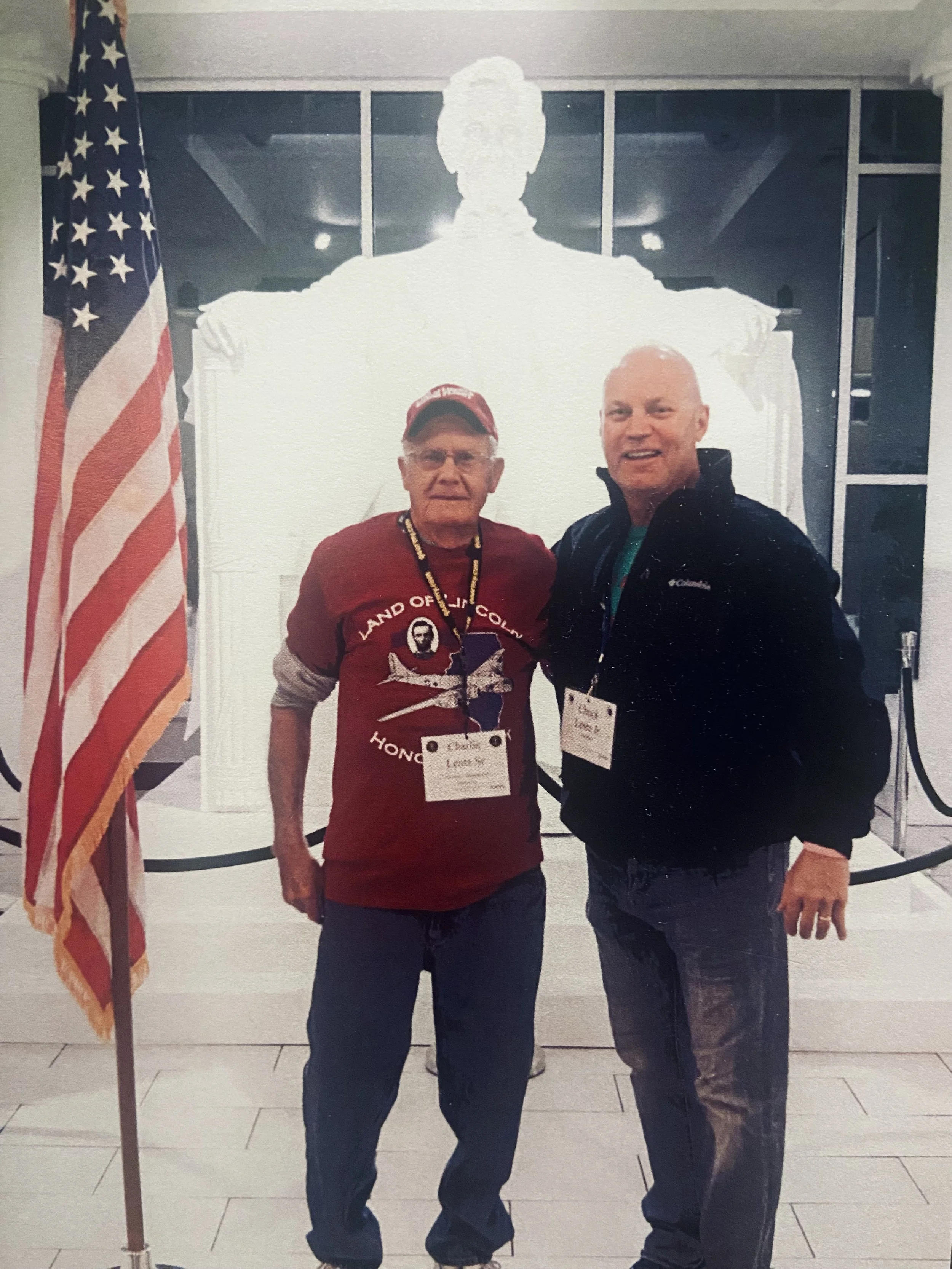 Two men standing indoors in front of an American flag and a large illuminated sculpture of a person. They are wearing name tags, and one is wearing a red shirt with patriotic designs.