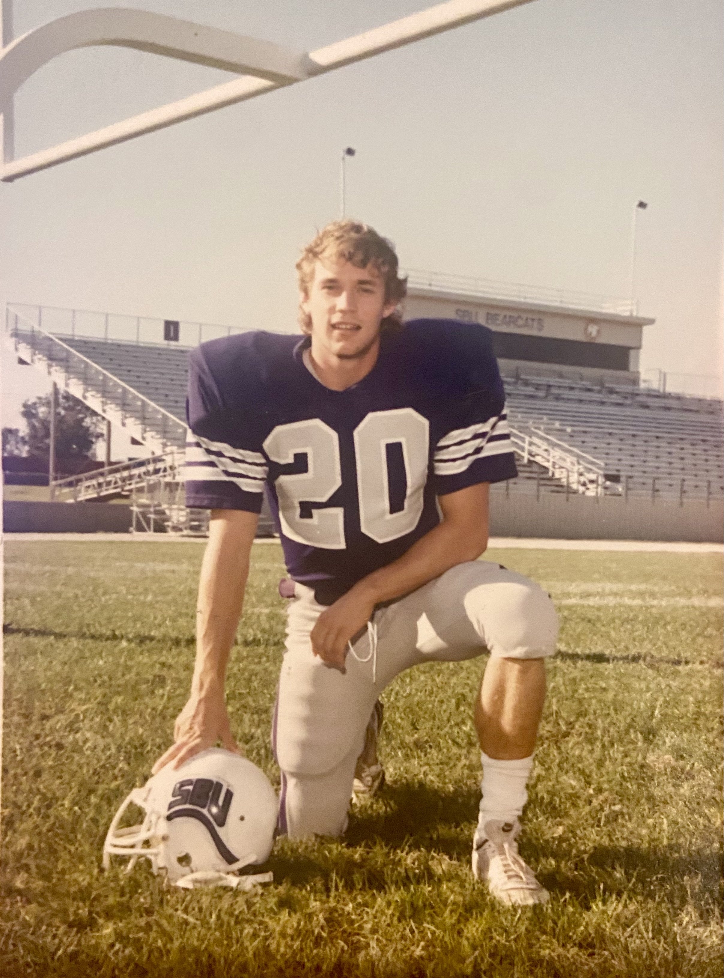 A young man in American football uniform kneeling on a football field, holding a white helmet with the letters 'SOU' and the number 20 on his jersey, with stadium bleachers in the background.