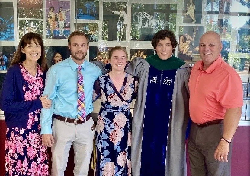 Family celebrating graduation, dressed in colorful clothing, with graduation cap and gown, indoors with photos on the wall