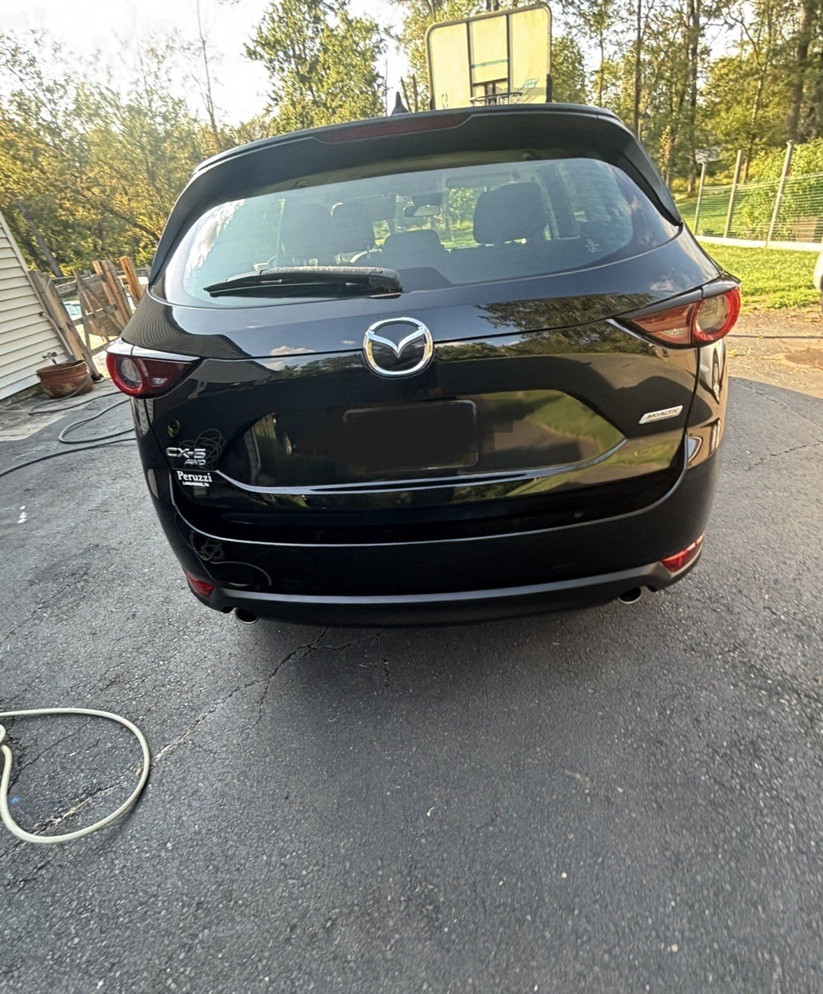 Rear view of a black Mazda CX-5 parked on a driveway, with trees and a fence in the background.