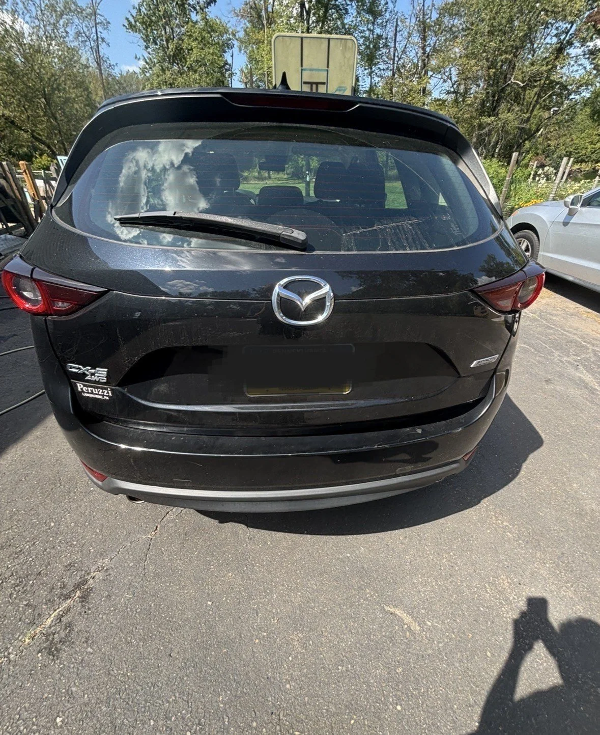 Rear view of a black Mazda CX-5 parked on a paved surface, with trees and a partly cloudy sky reflected in the rear window.