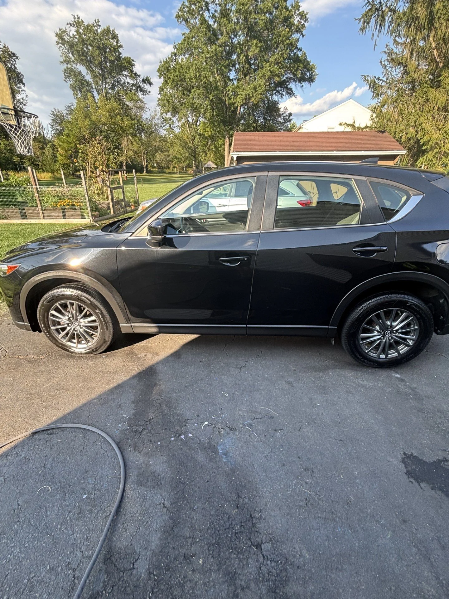 Black SUV parked on cracked asphalt driveway with grass yard, trees, and a house in the background.
