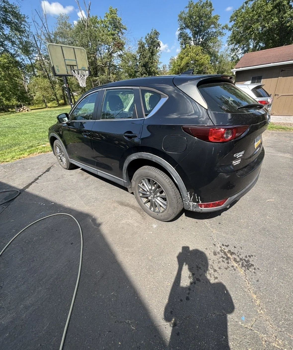 Black Mazda SUV parked on a driveway near a house with a basketball hoop in a residential area, with trees and a blue sky in the background.