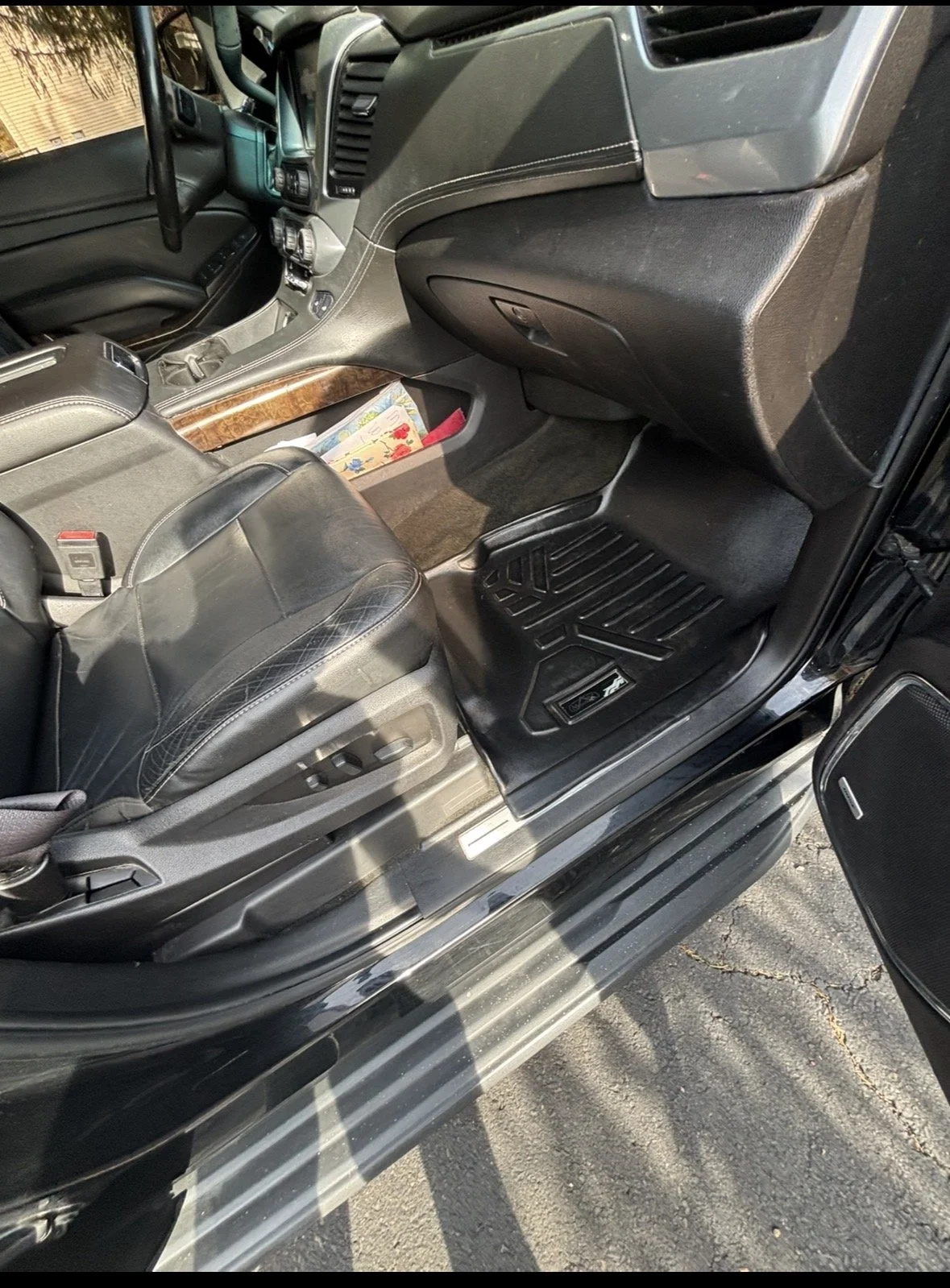 The interior of a vehicle shows the front passenger seat, dashboard, and a small storage area with a floral-patterned book. The vehicle has black leather seats and a wood-trim accent on the center console.