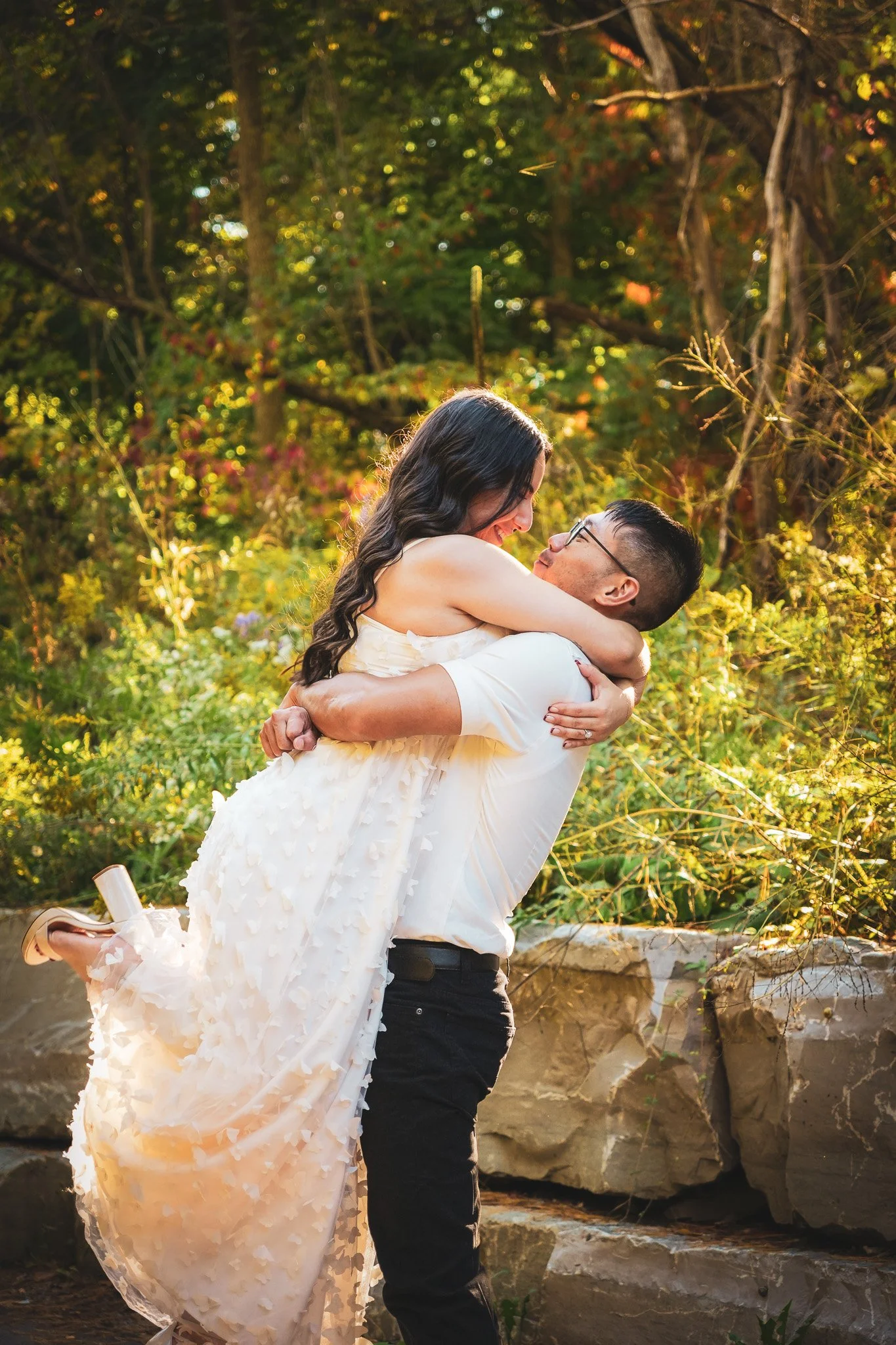 A man lifting a woman in a white dress in a forested area with sunlight filtering through trees.