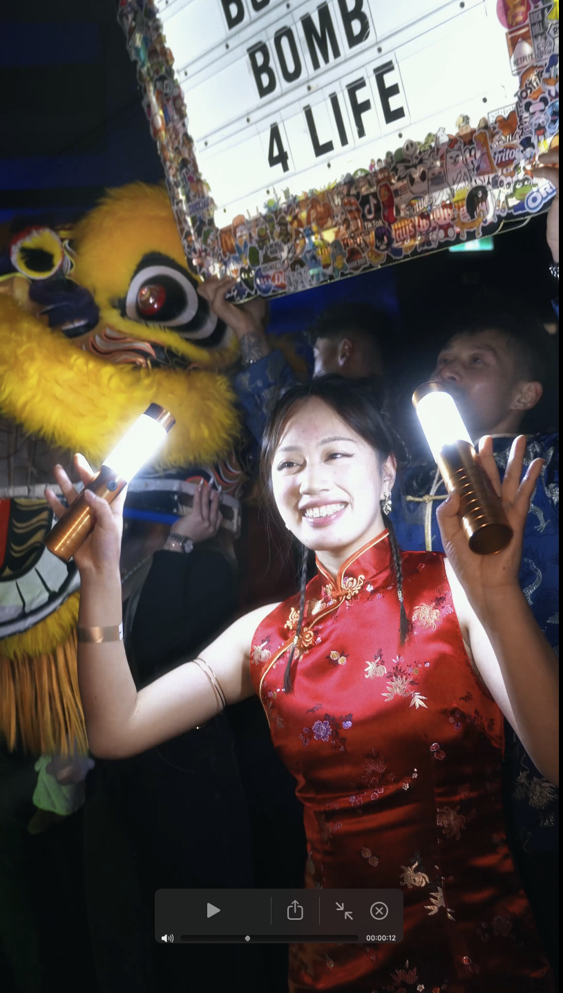 A woman in a traditional red Chinese dress smiling and holding two glow sticks at a celebration. Behind her, a person in a yellow lion costume and a sign that reads 'BOMB 4 LIFE' with numerous stickers.