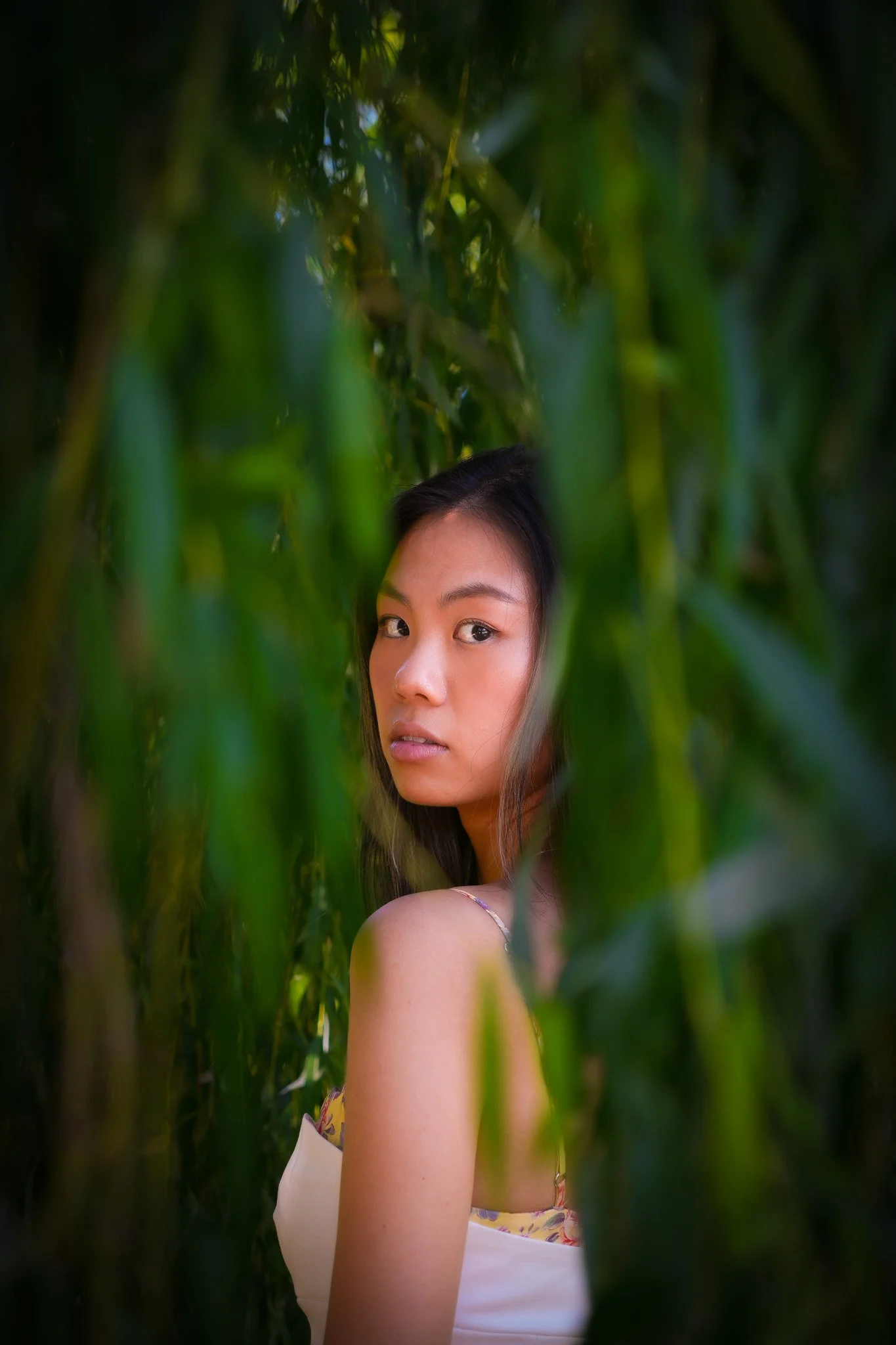 A young woman with dark hair and a serious expression, looking over her shoulder, surrounded by tall green plants or tall grass.