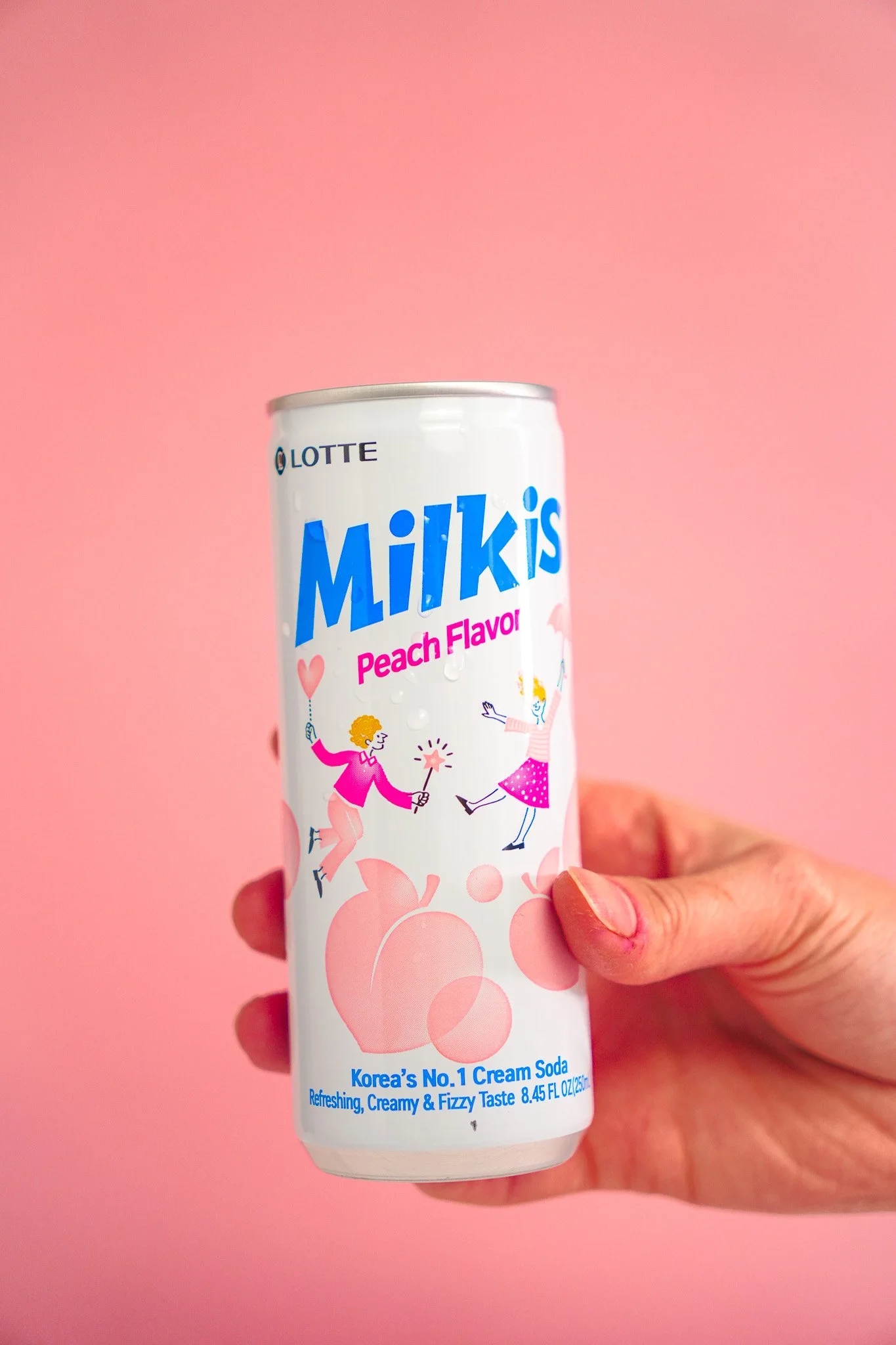 Hand holding a can of Milkis Peach Flavor cream soda against a pink background.