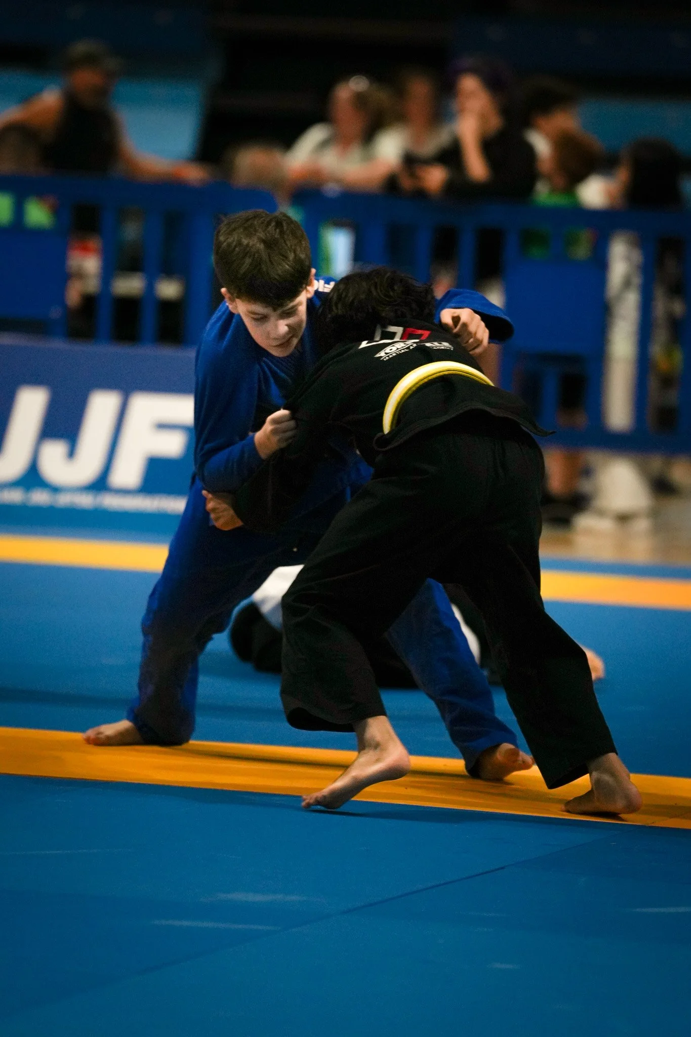 Two young boys compete in a Brazilian Jiu-Jitsu match on a blue mat, with a yellow line, surrounded by spectators.