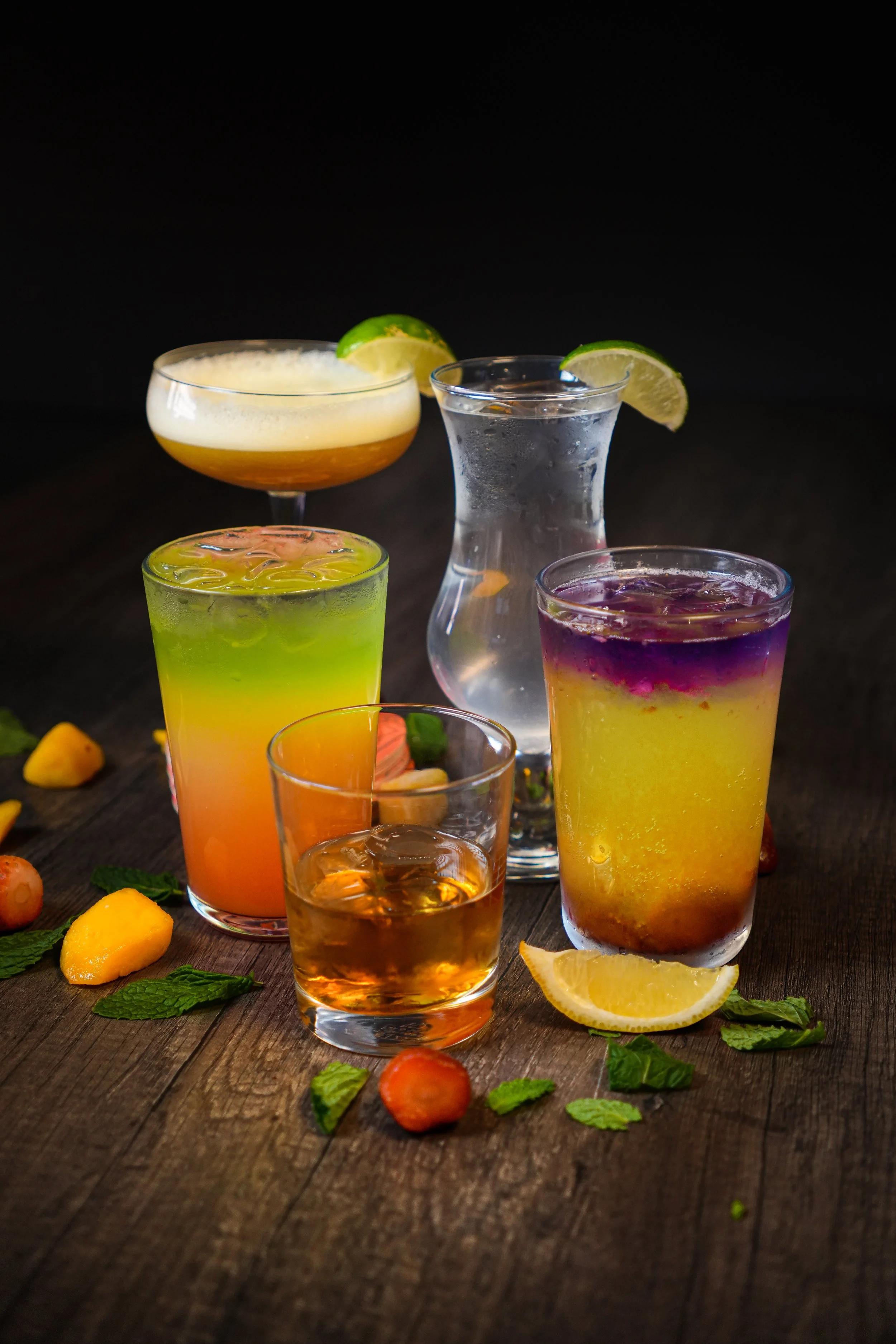 Assorted colorful cocktails with lime and lemon slices, garnished with mint leaves, on a rustic wooden table.