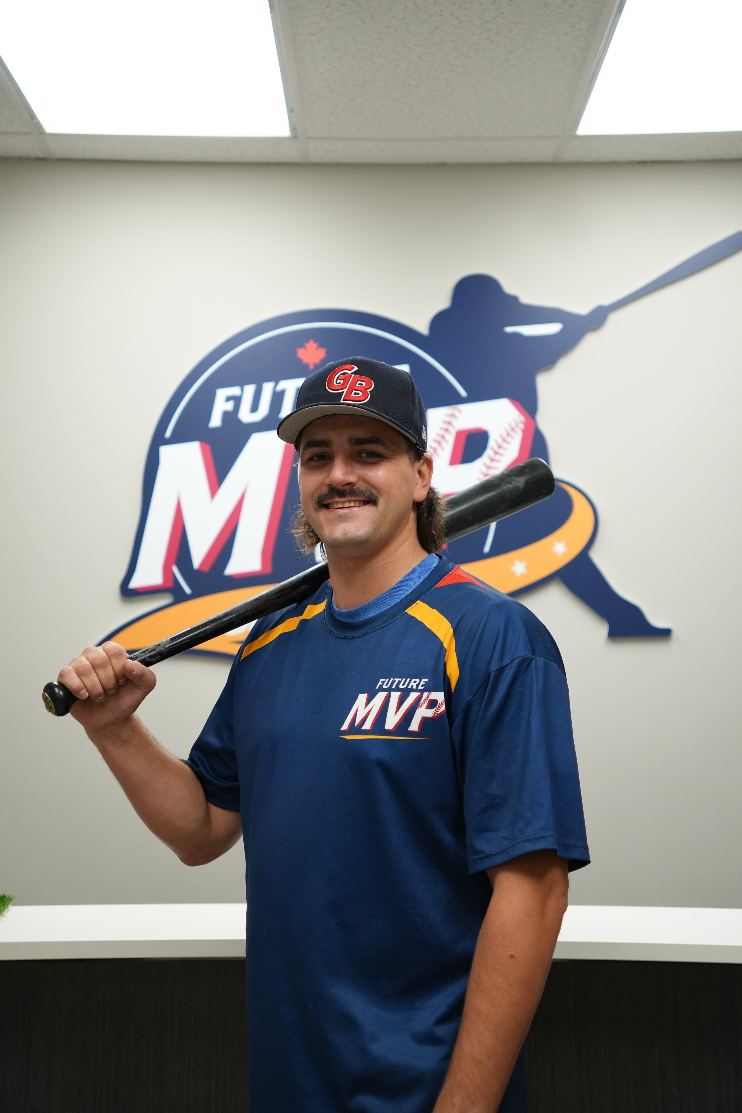 Young man wearing a blue Future MVP baseball shirt and a blue cap with red and white letters, holding a baseball bat over his shoulder, standing in front of a wall with a sports-themed logo and silhouette of a baseball player.