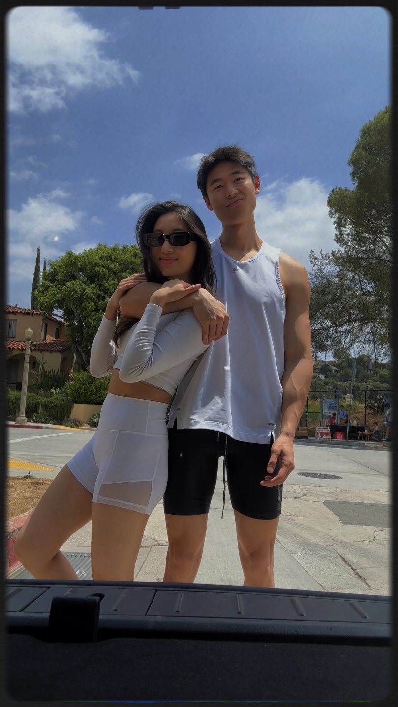 Two young people, a woman and a man, posing outdoors on a sunny day, with trees and residential buildings in the background.