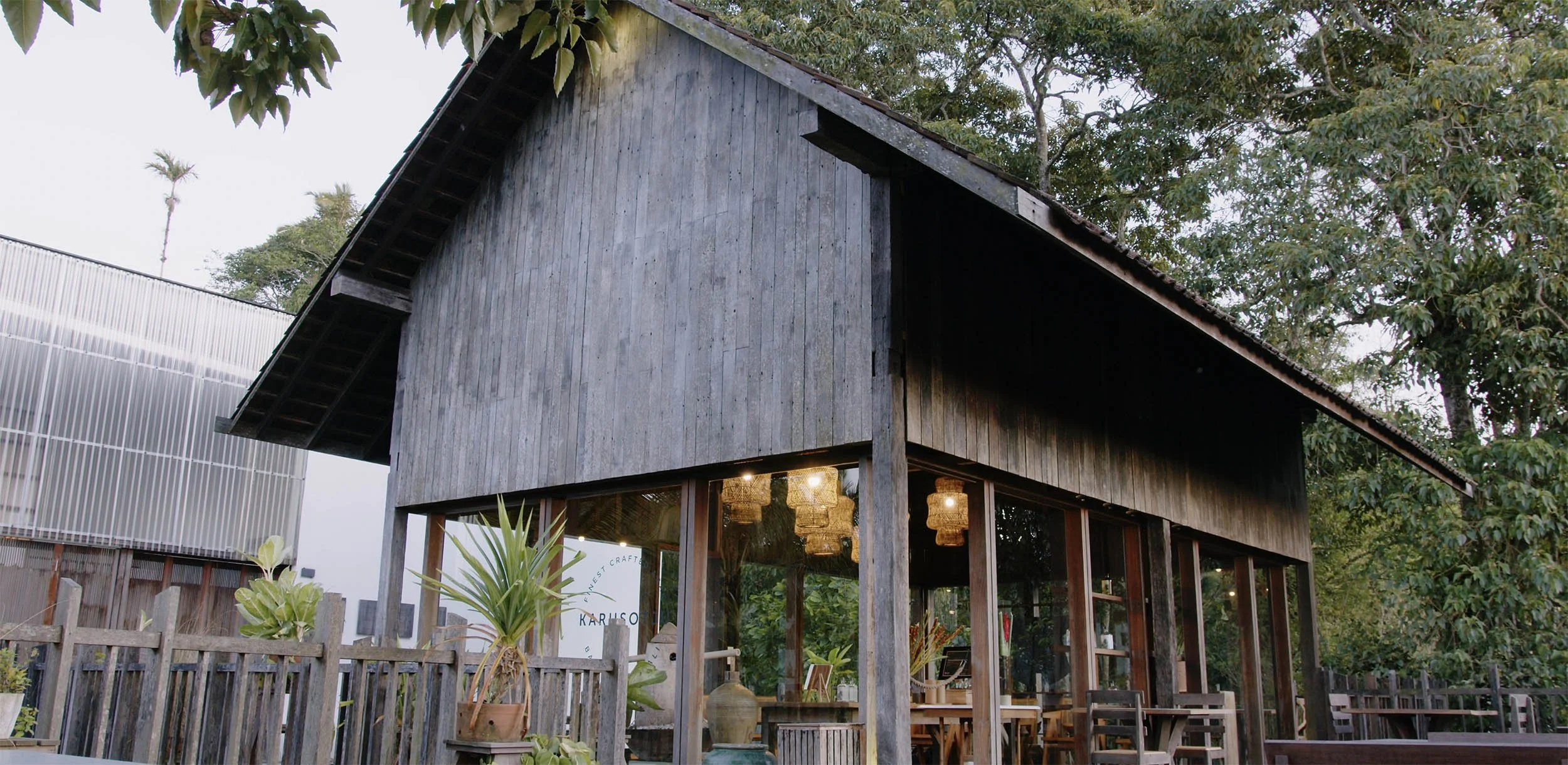 A two-story wooden building with a large glass front, surrounded by outdoor seating with wooden tables and chairs, and a garden with potted plants, set in a lush green environment.