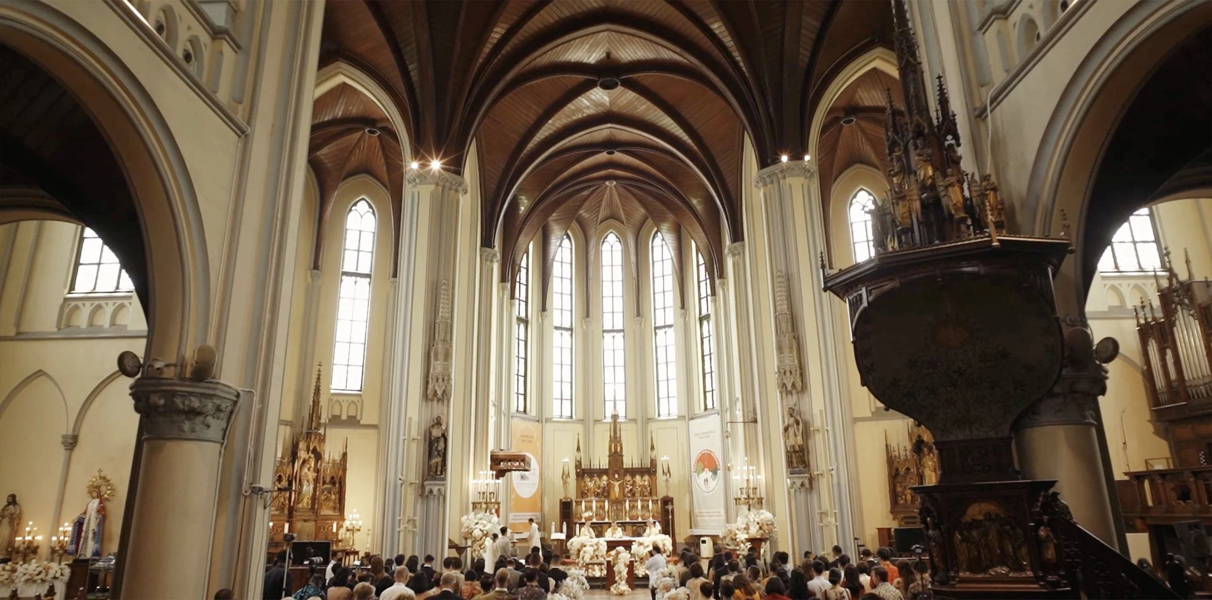 Interior of a church with high pointed arched windows, wooden ceiling, altar decorated with flowers, and a large congregation attending a religious service.