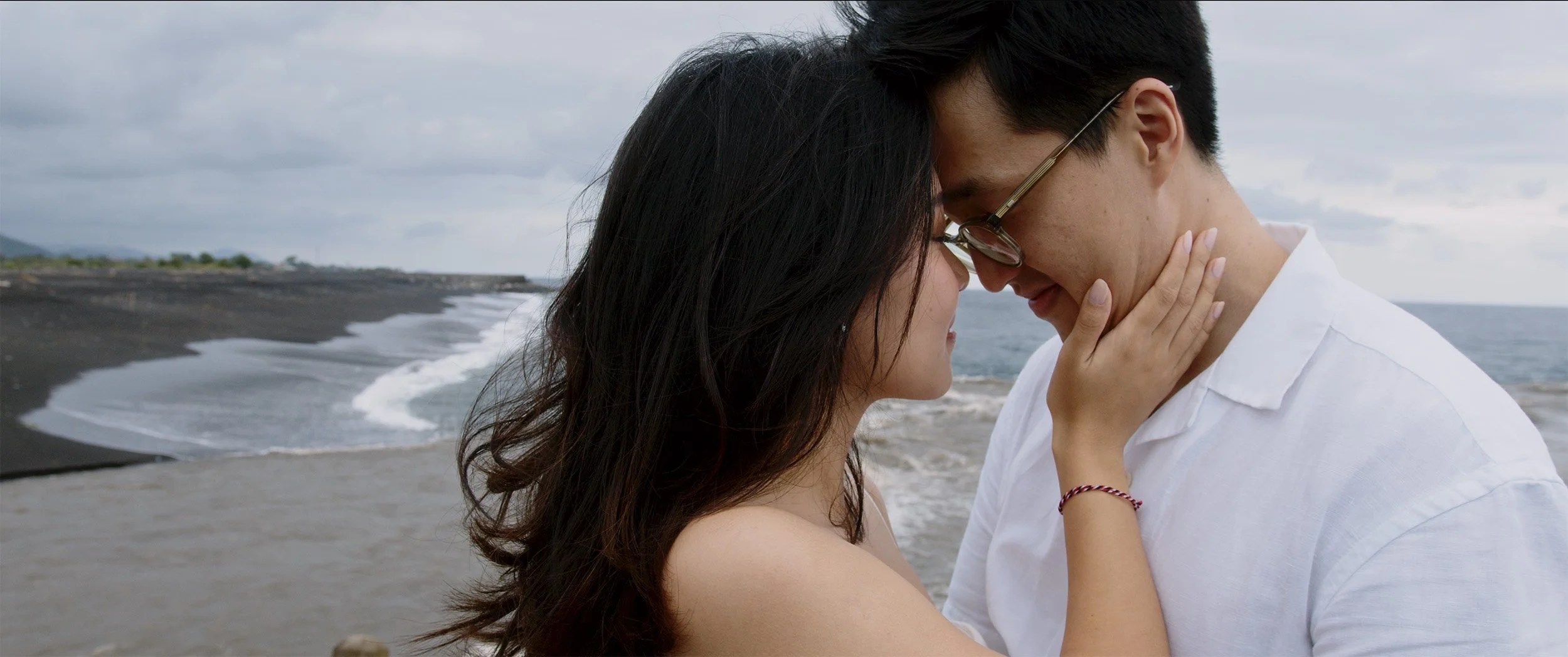 A couple with foreheads touching and eyes closed on the beach, with waves and cloudy sky in the background.