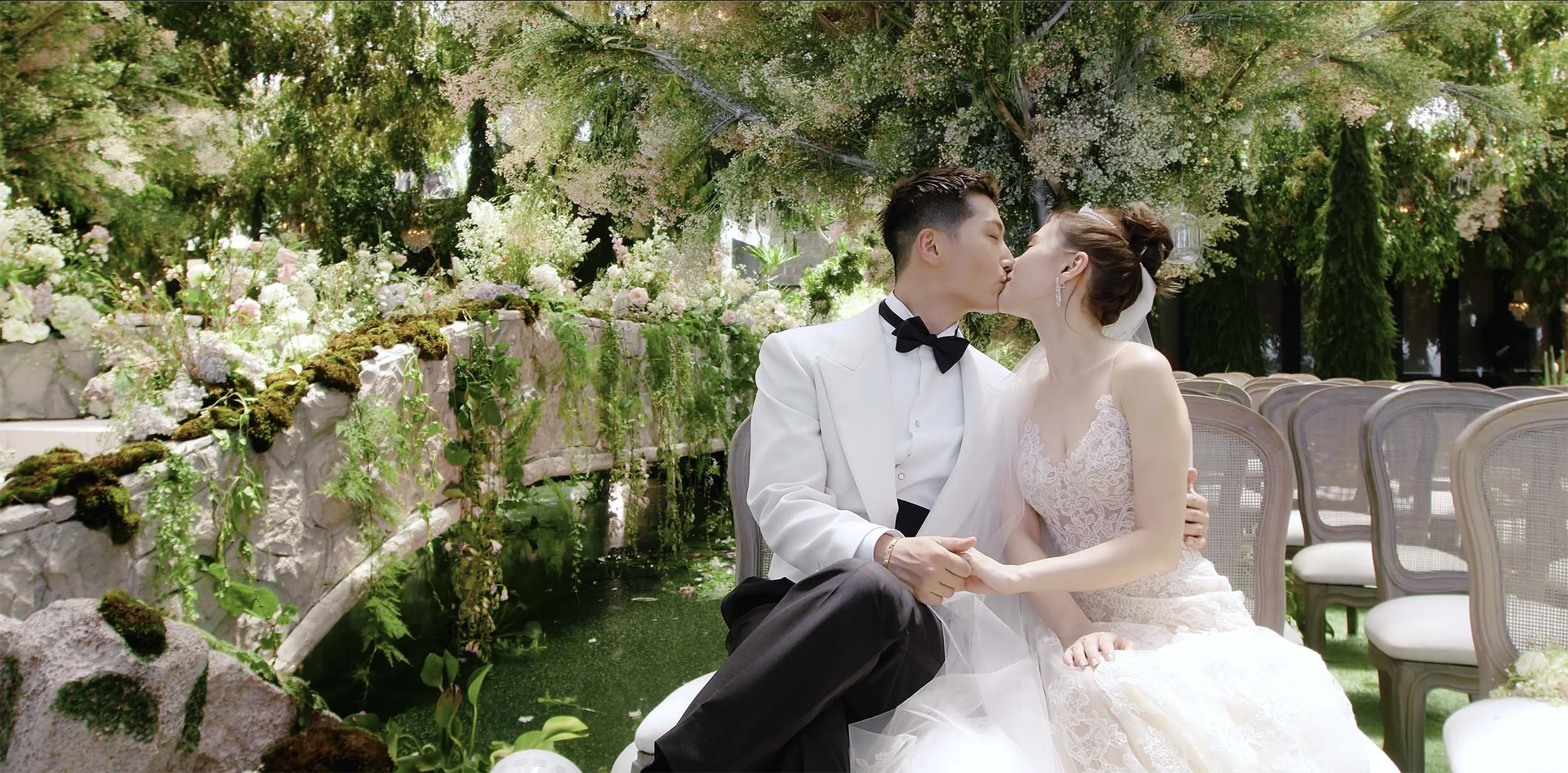 A newlywed couple in wedding attire sharing a kiss outdoors amid greenery, flowers, and trees, seated on white chairs.