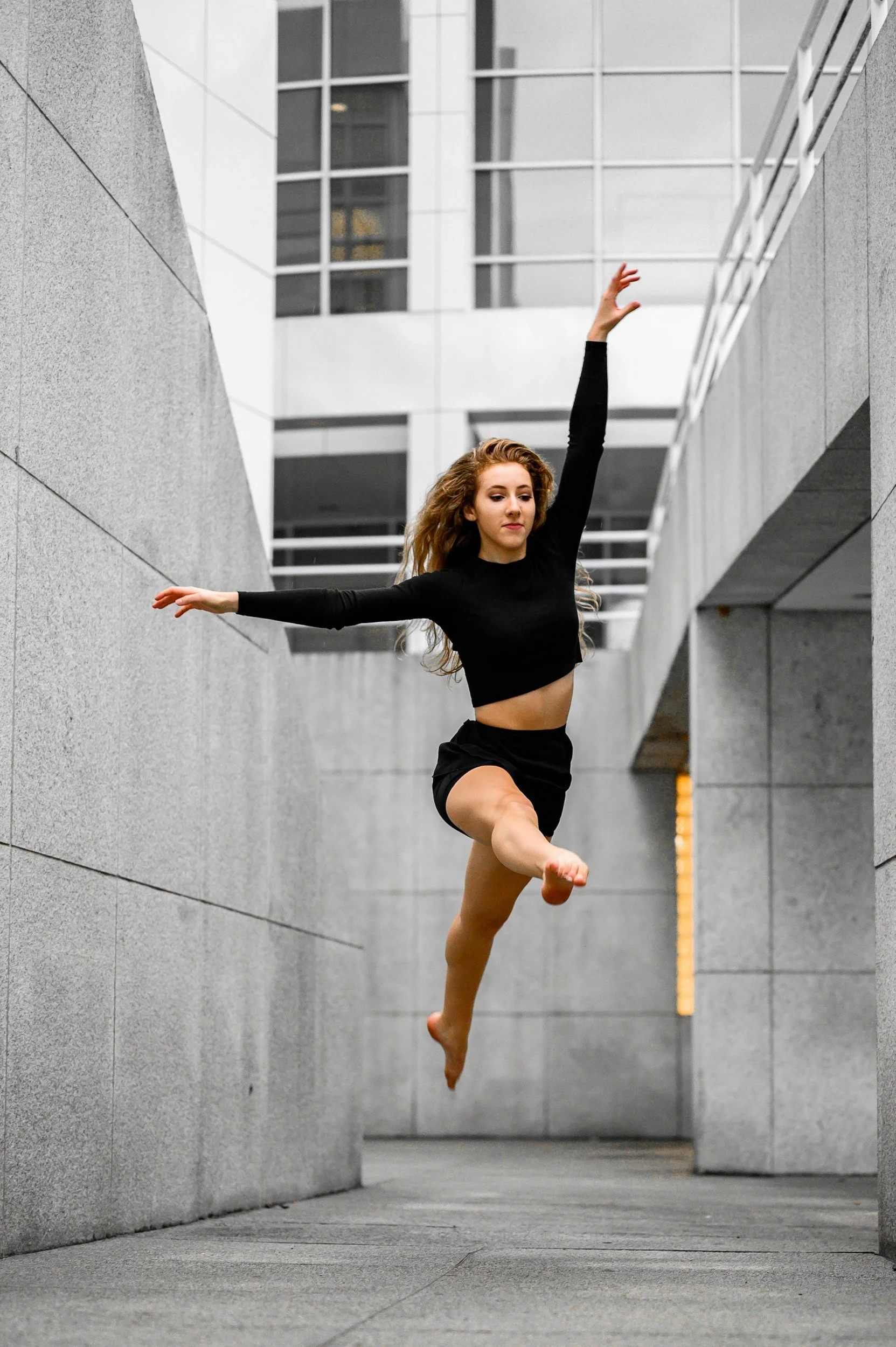 A young woman in black athletic clothing performing a leap in an urban outdoor setting with modern gray buildings and concrete walkways.