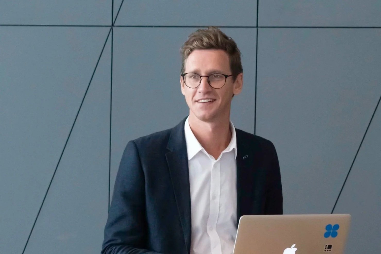 A man with glasses, wearing a dark blazer and white shirt, standing in front of a gray wall with geometric lines, holding a laptop.