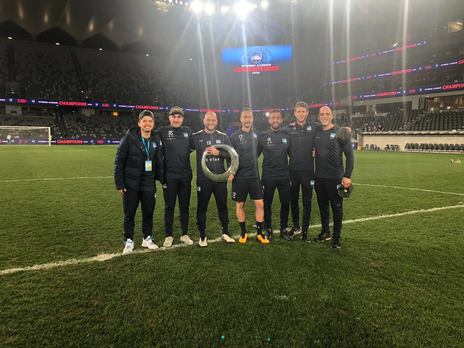Group of seven soccer players and staff members standing on a soccer field celebrating with a trophy after a victory, with empty stadium seats and a scoreboard in the background.