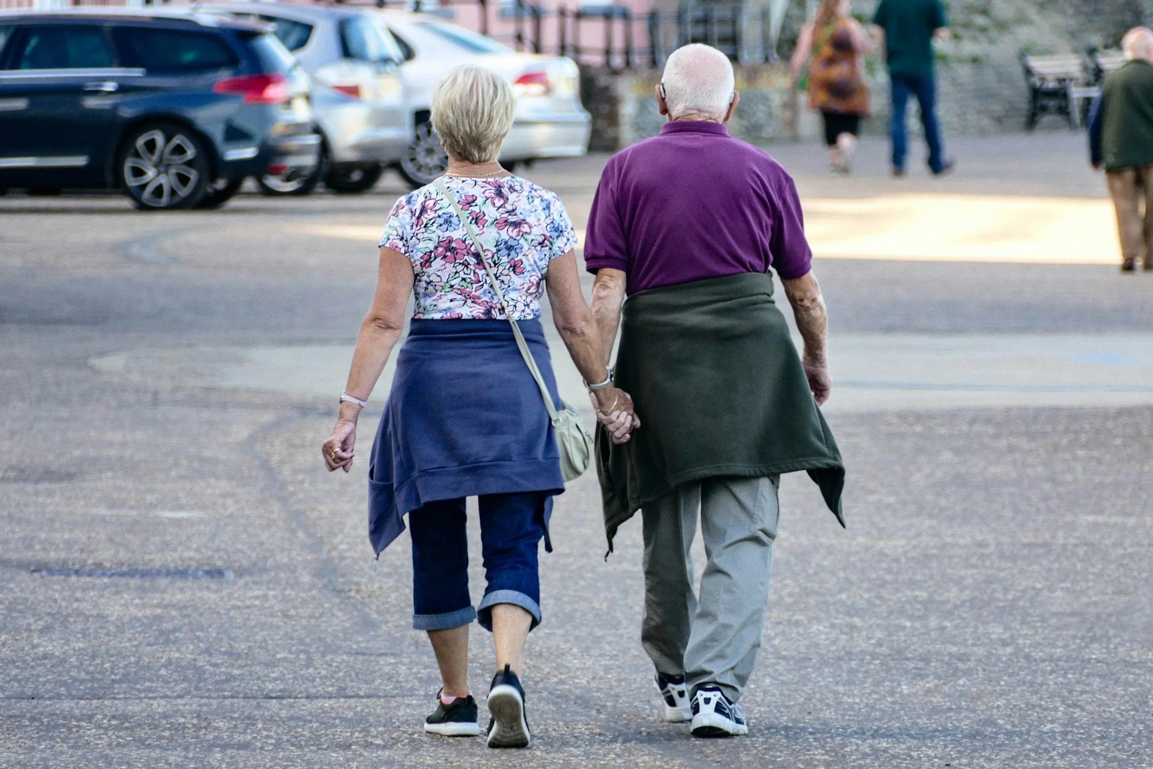Two elderly women are walking together hand-in-hand across a street, seen from behind. One wears a colorful floral top with rolled-up jeans and sneakers, while the other wears a purple shirt with gray pants and sneakers. Cars are parked along the street in the background.
