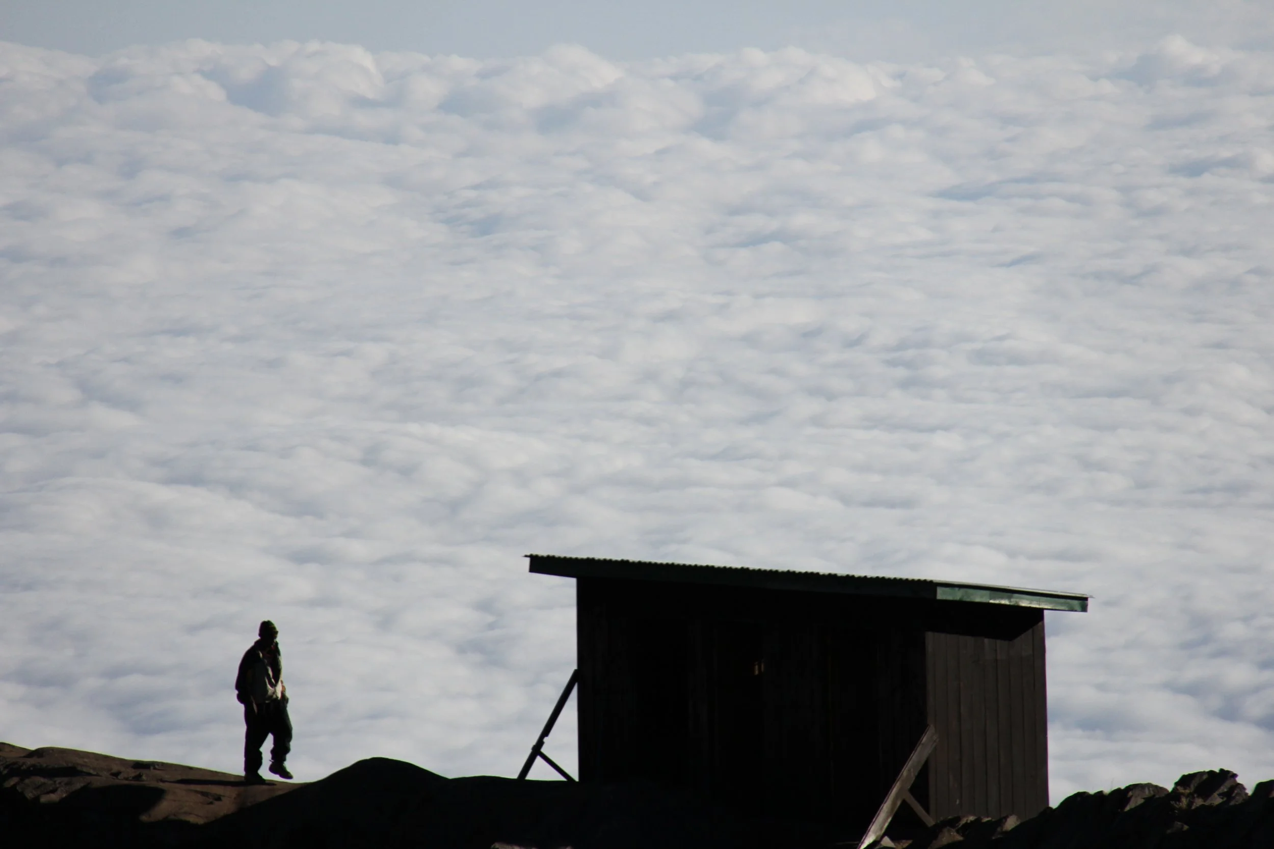 Silhouette of a person standing on rocky ground near a small, dark wooden structure, with a cloudy sky in the background.