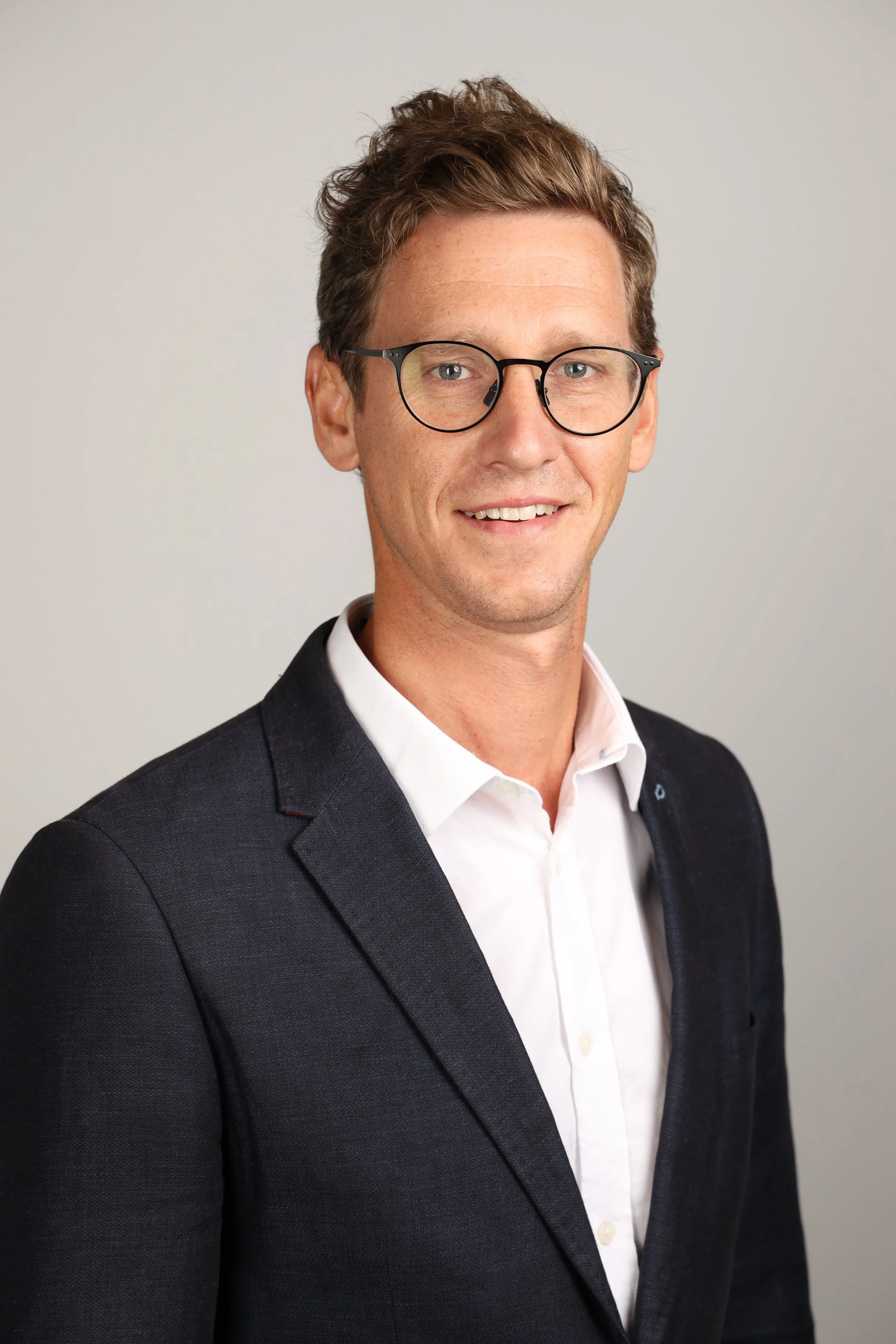 Headshot of a man with short, wavy brown hair, wearing black-rimmed glasses, a white dress shirt, and a dark suit jacket, smiling slightly against a plain light gray background.