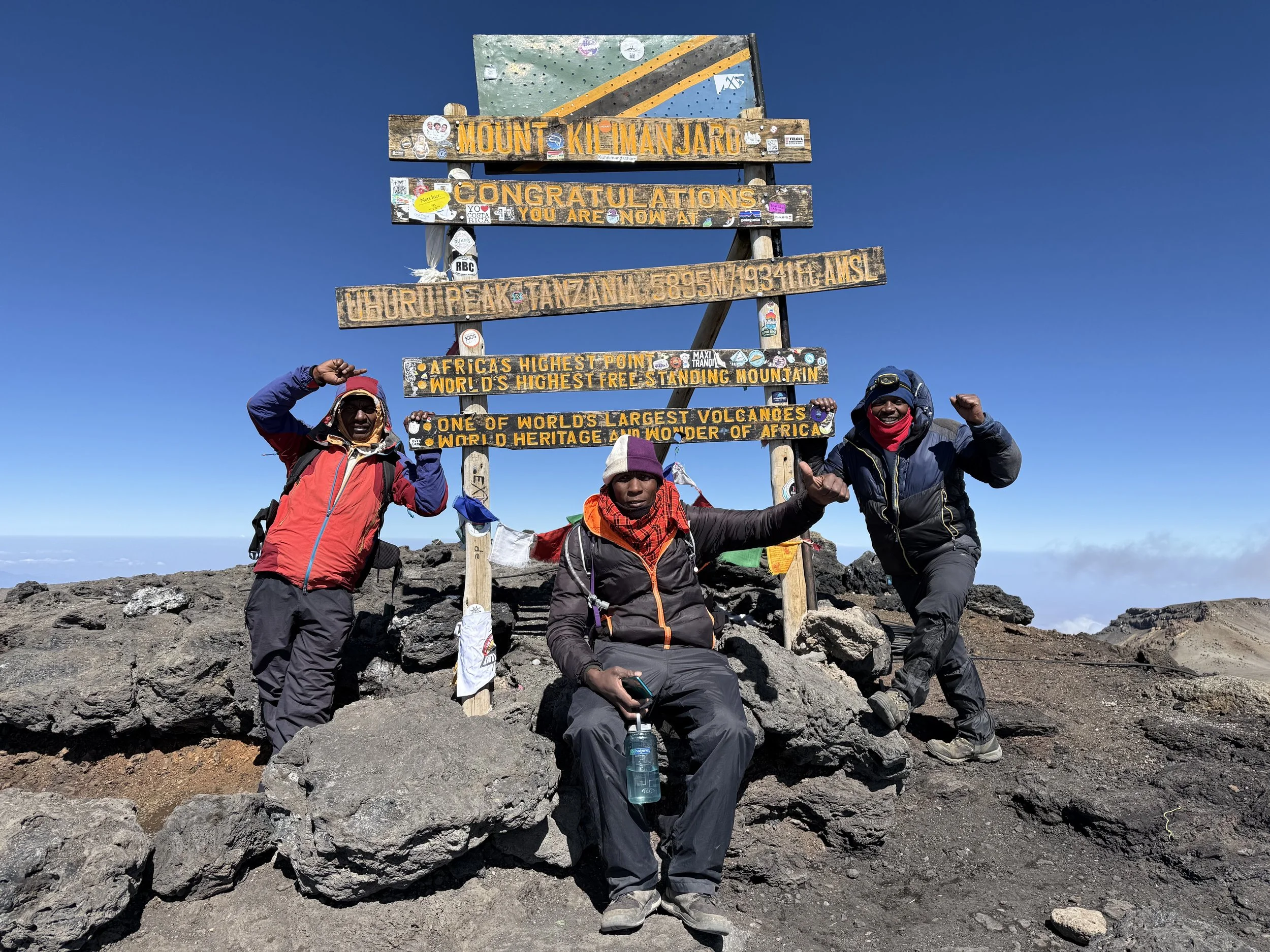 Three climbers at the summit of Mount Kilimanjaro, celebrating with arms raised, standing in front of a sign that reads 'Mount Kilimanjaro, Tanzania, 5895m, 19341 ft AMSL'. The sign also announces it as Africa's highest point and the world's highest free-standing mountain, with other details about the volcano's size and heritage. The climbers are dressed in outdoor gear suitable for cold and windy conditions, with a clear blue sky above and rocky terrain around them.