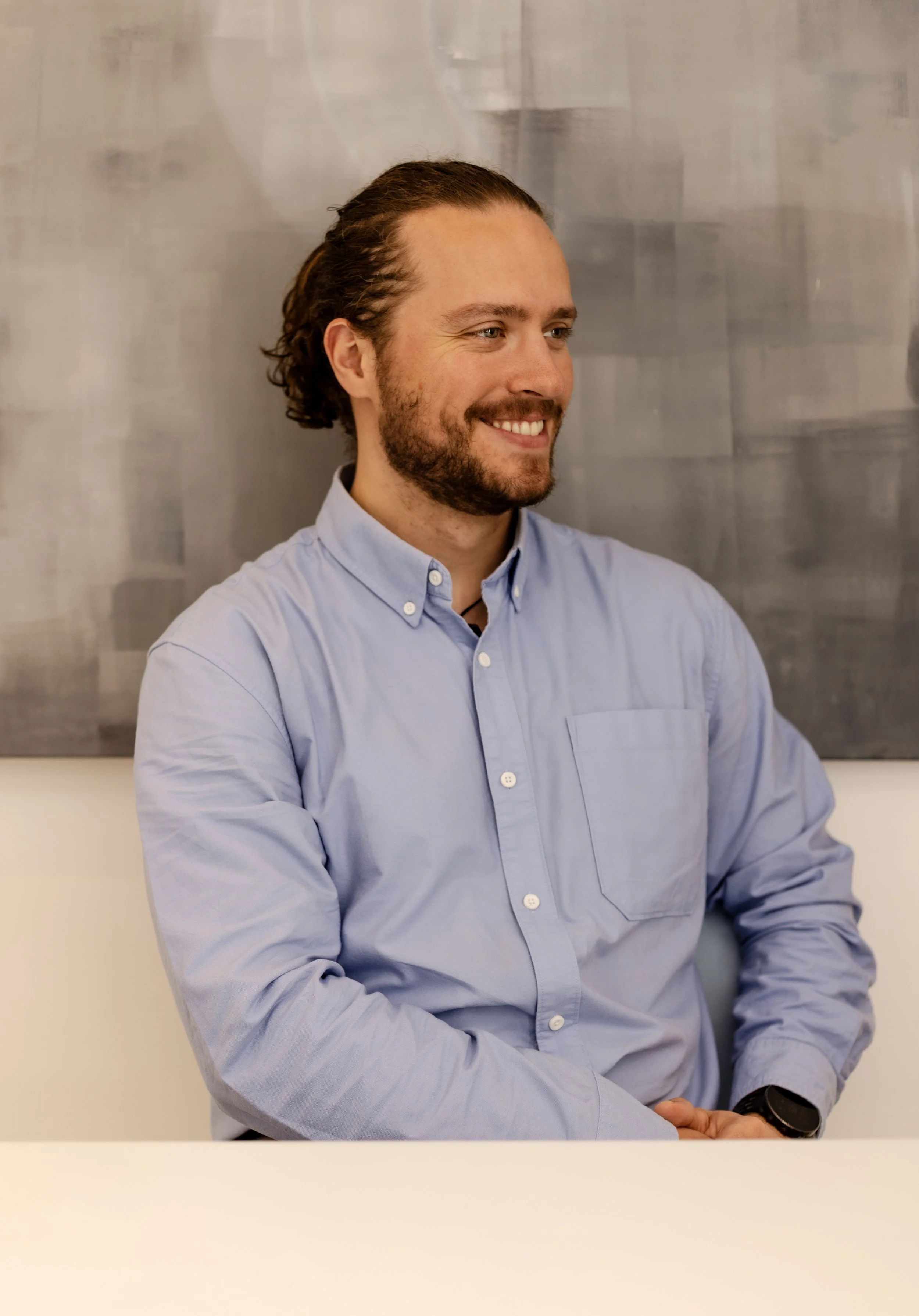 Portrait of a executive coach Greg Chambers. Beard and long hair, wearing a light blue button-down shirt against a grey background smiling and looking to the side.